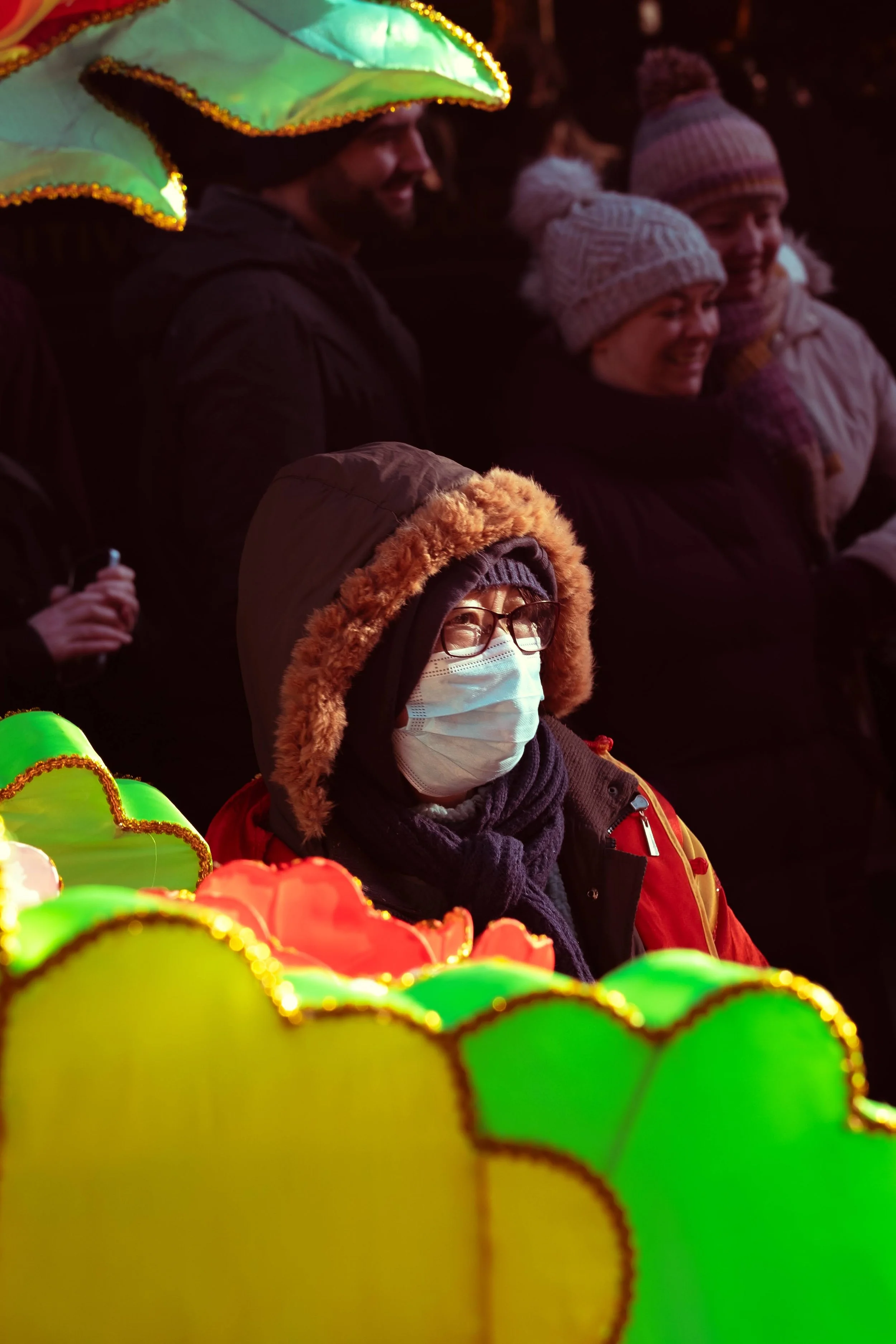 A group of people at night, dressed warmly in hats, scarves, and coats. One person in the foreground is wearing glasses and a face mask with a hood and fur lining, standing near brightly colored, illuminated lanterns.