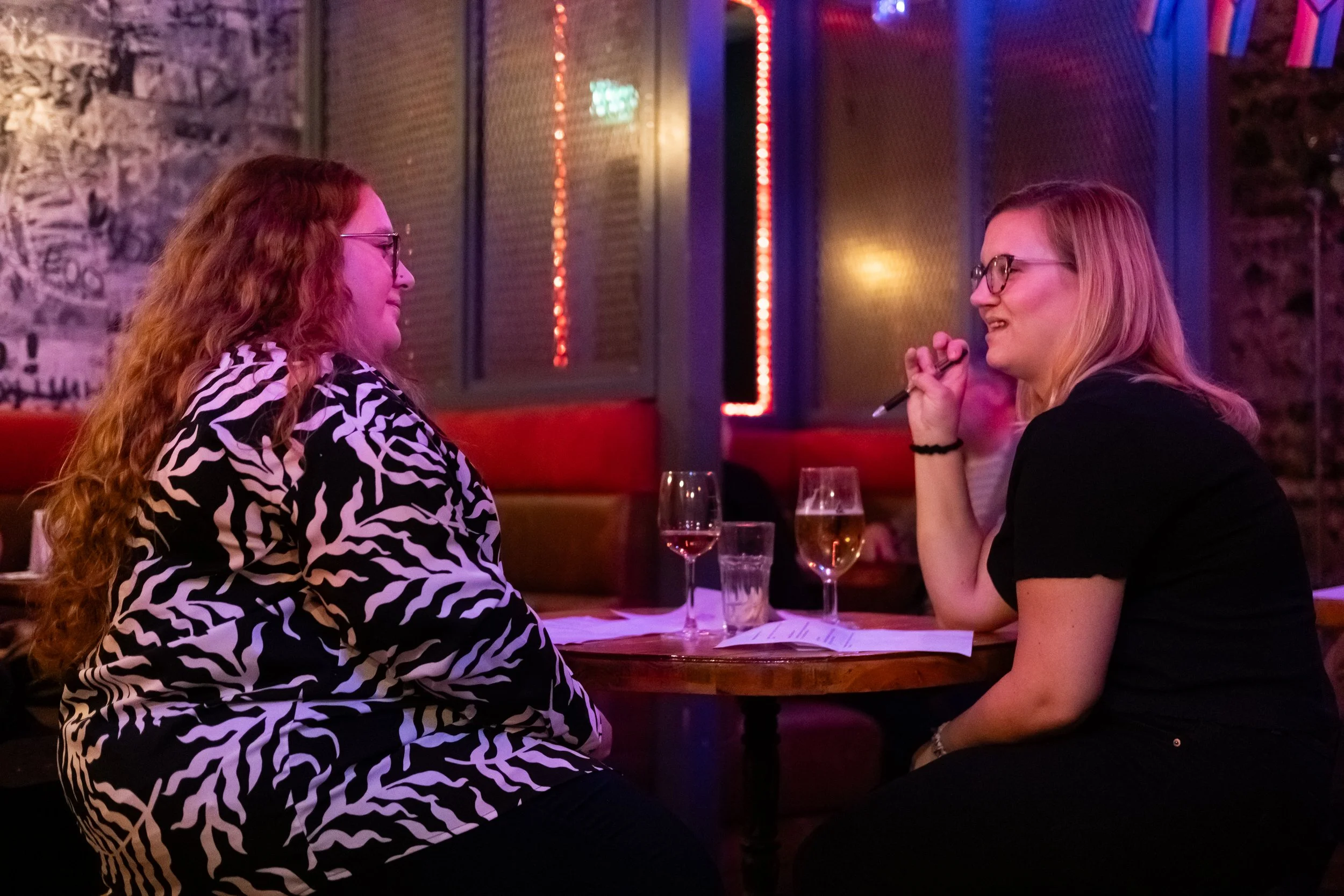Two women sitting at a table having a conversation in a dimly lit bar or restaurant with neon lights, glasses of drinks, and casual atmosphere.