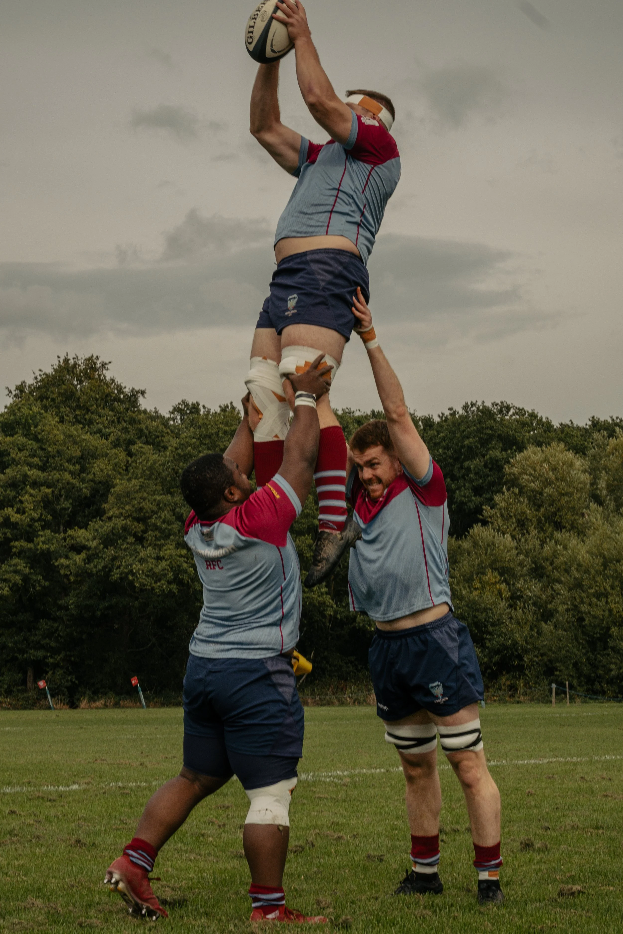 Three rugby players forming a human pyramid on a field, with a player at the top holding a rugby ball.
