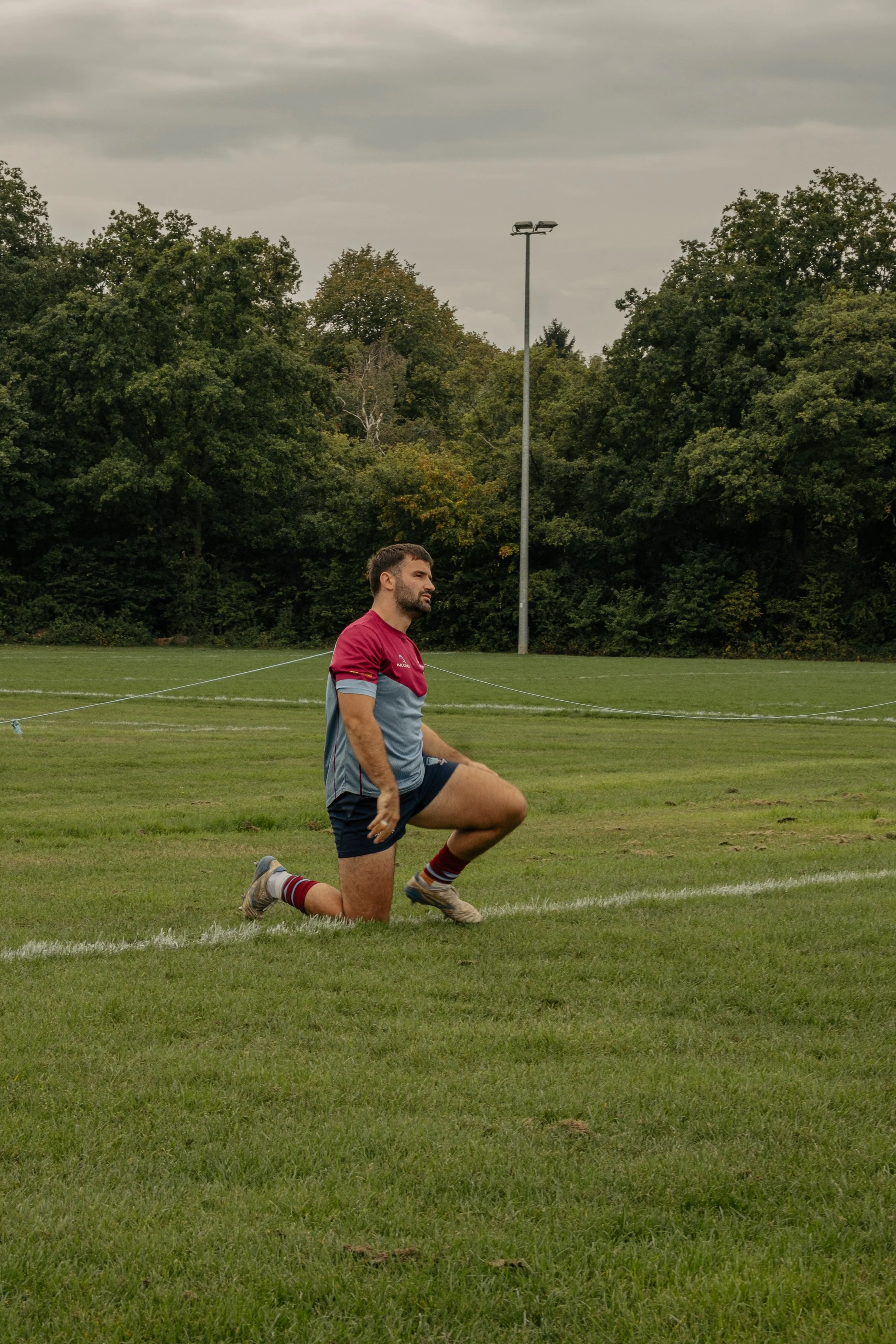 A man in athletic gear kneeling on one knee on a grassy sports field, with trees and cloudy sky in the background.