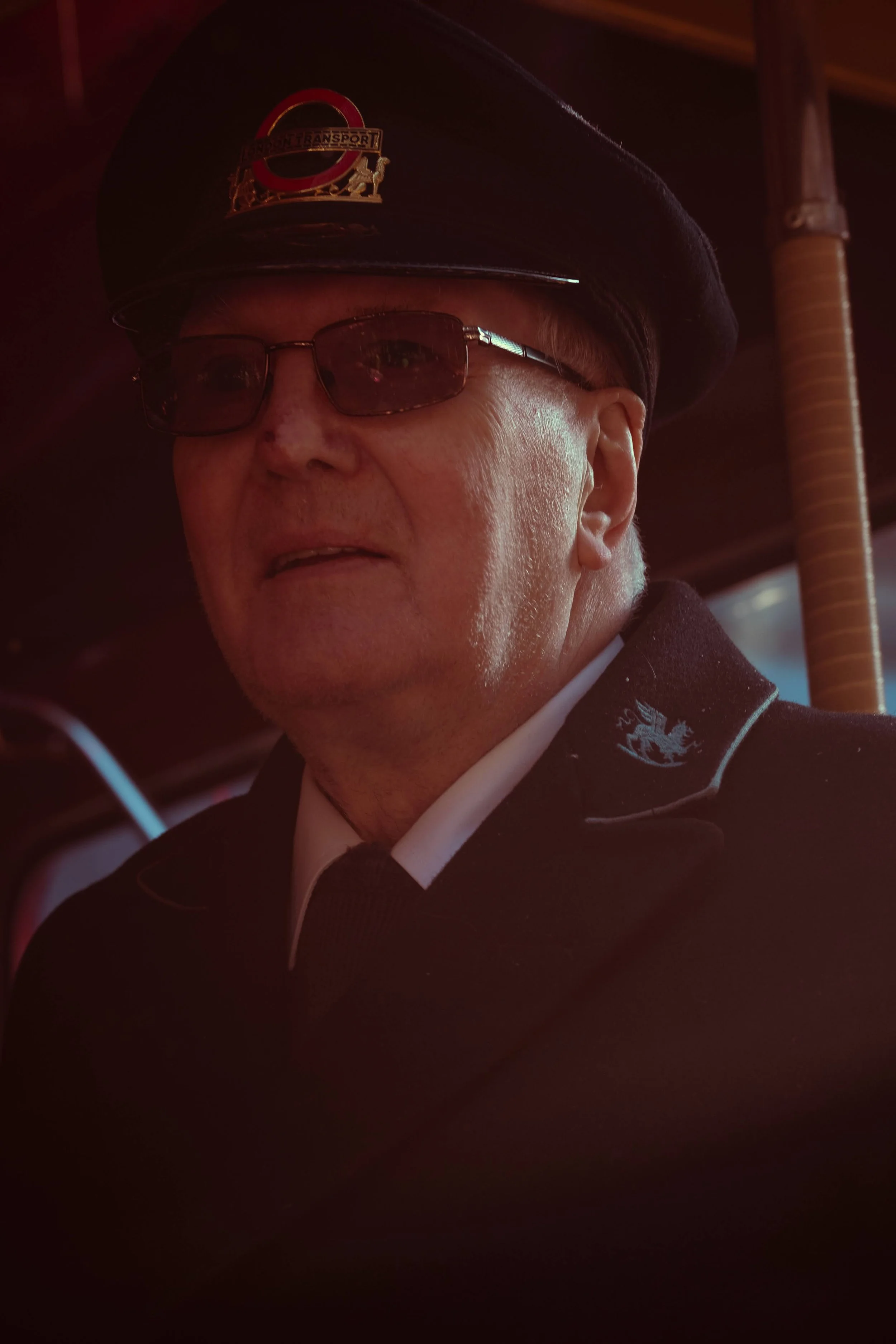 A man in a pilot uniform wearing a navy cap with a Transport for London insignia, sunglasses, and a dark suit, standing inside a vehicle.