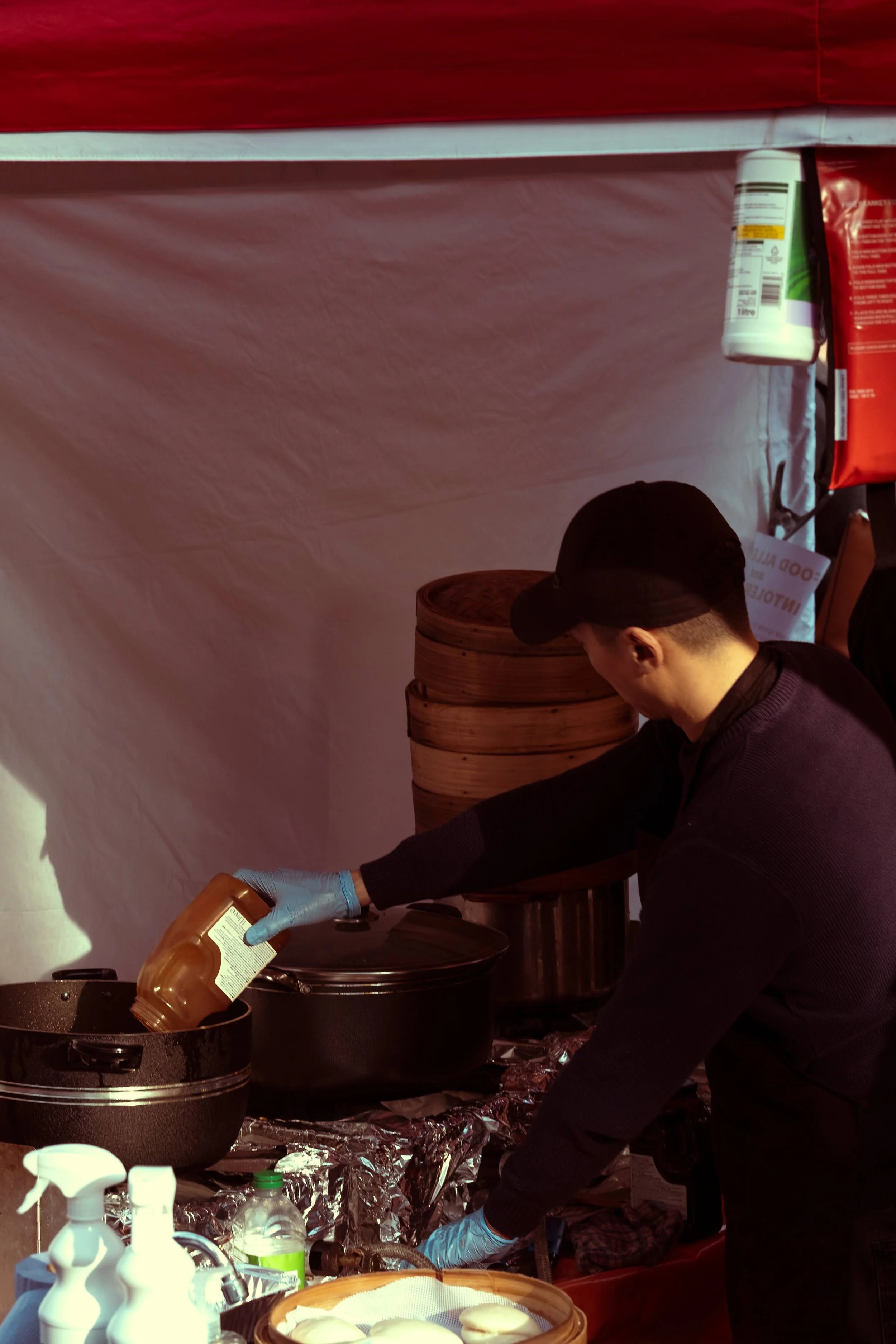 A person wearing a black cap, black sweater, and blue gloves pouring sauce into a pot at a food stall. There are stacked bamboo steamers and various condiments and utensils on the table.