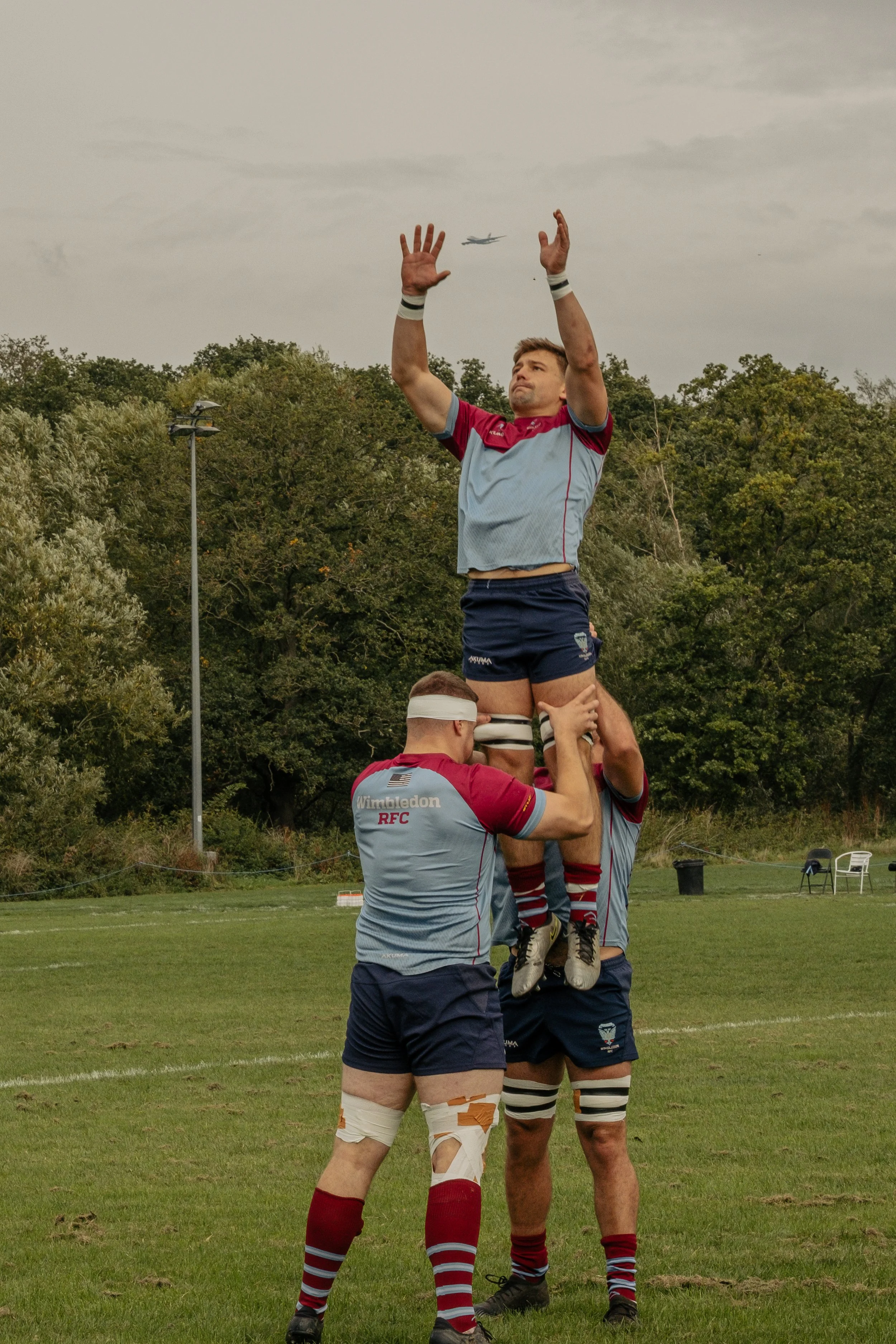 Rugby players performing a line-out on a grassy field during daytime, with one player being lifted by two teammates while another teammate stands nearby, with trees and a cloudy sky in the background.
