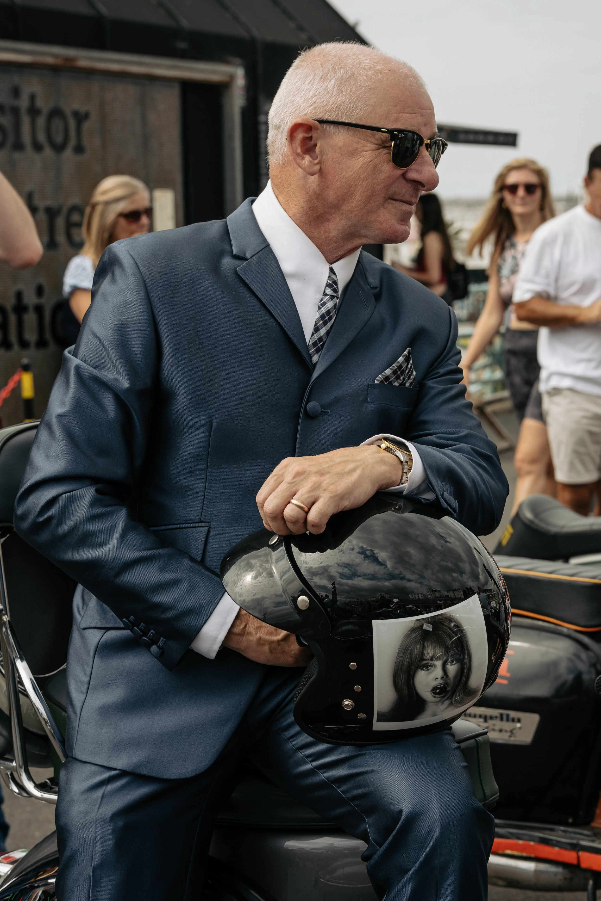 A man in a blue suit and sunglasses holding a black motorcycle helmet with a black and white portrait of a woman on it, sitting on a motorcycle at an outdoor event with people in the background.