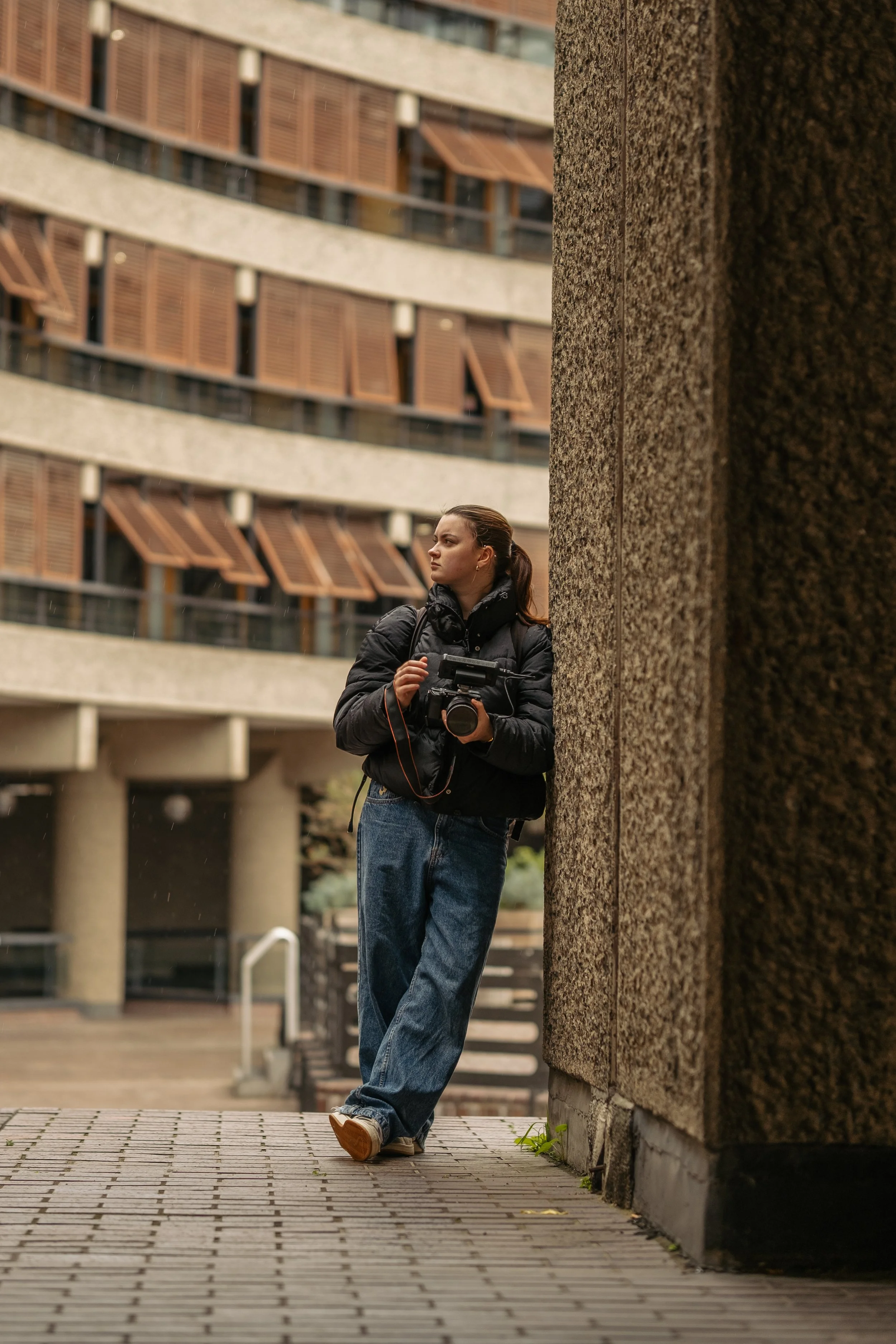 A woman with a camera and backpack leaning against a concrete wall outdoors, with a modern building in the background.