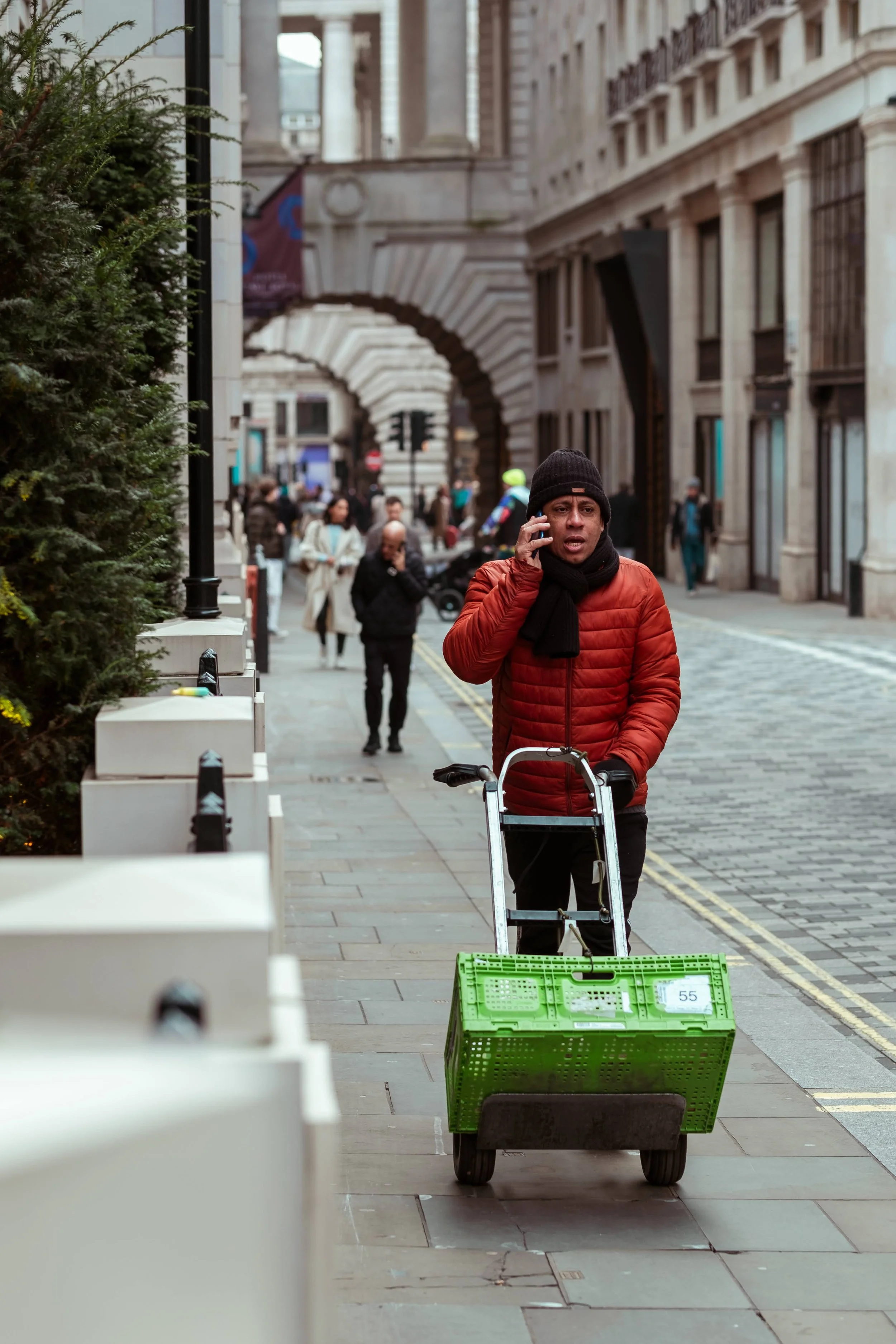 A man in a red jacket and black beanie talking on a cellphone while pulling a green shopping cart along a city sidewalk, with several pedestrians and historic buildings in the background.