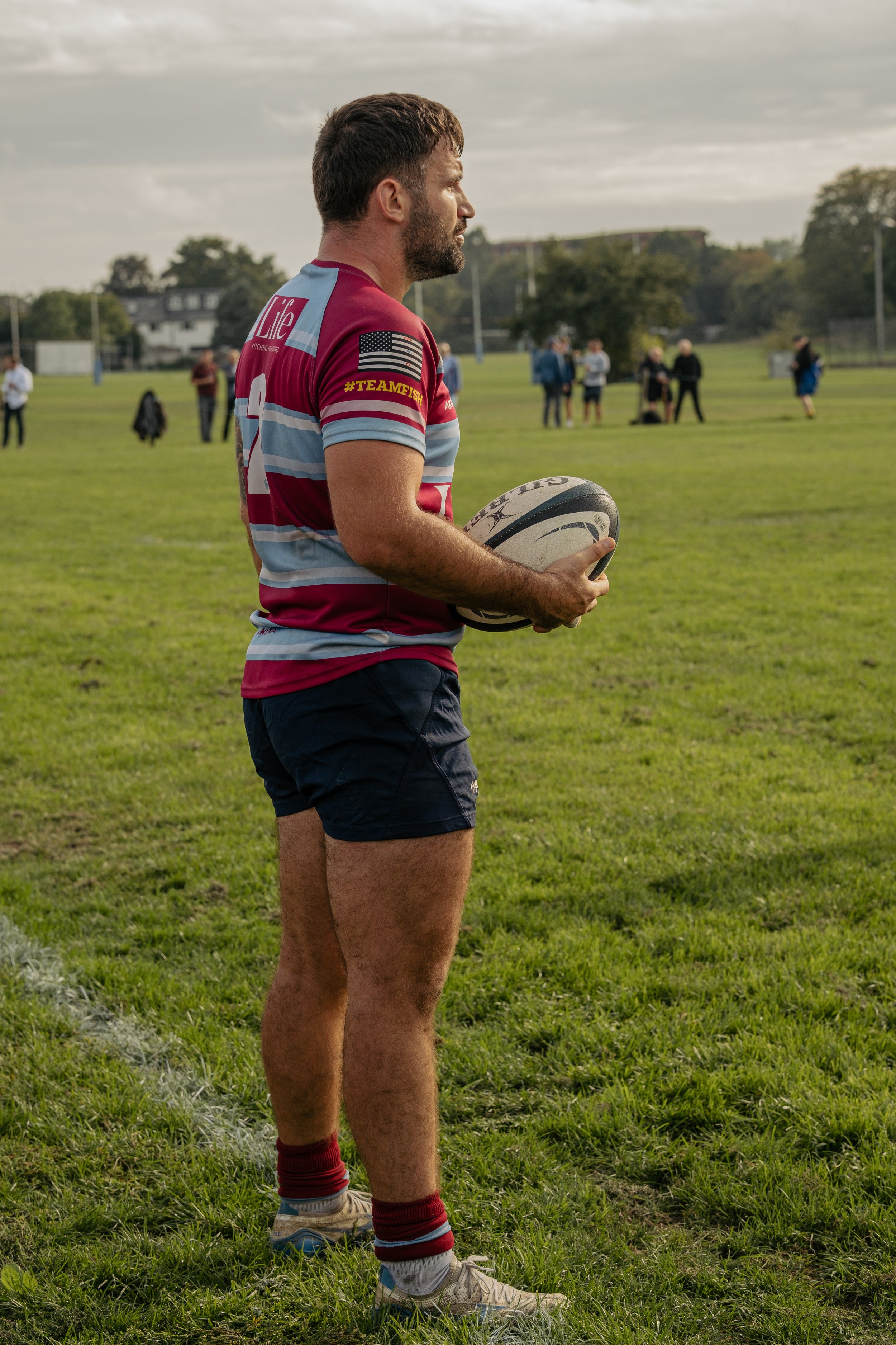 A man in a rugby uniform holding a rugby ball on a grassy field, with people in the background.