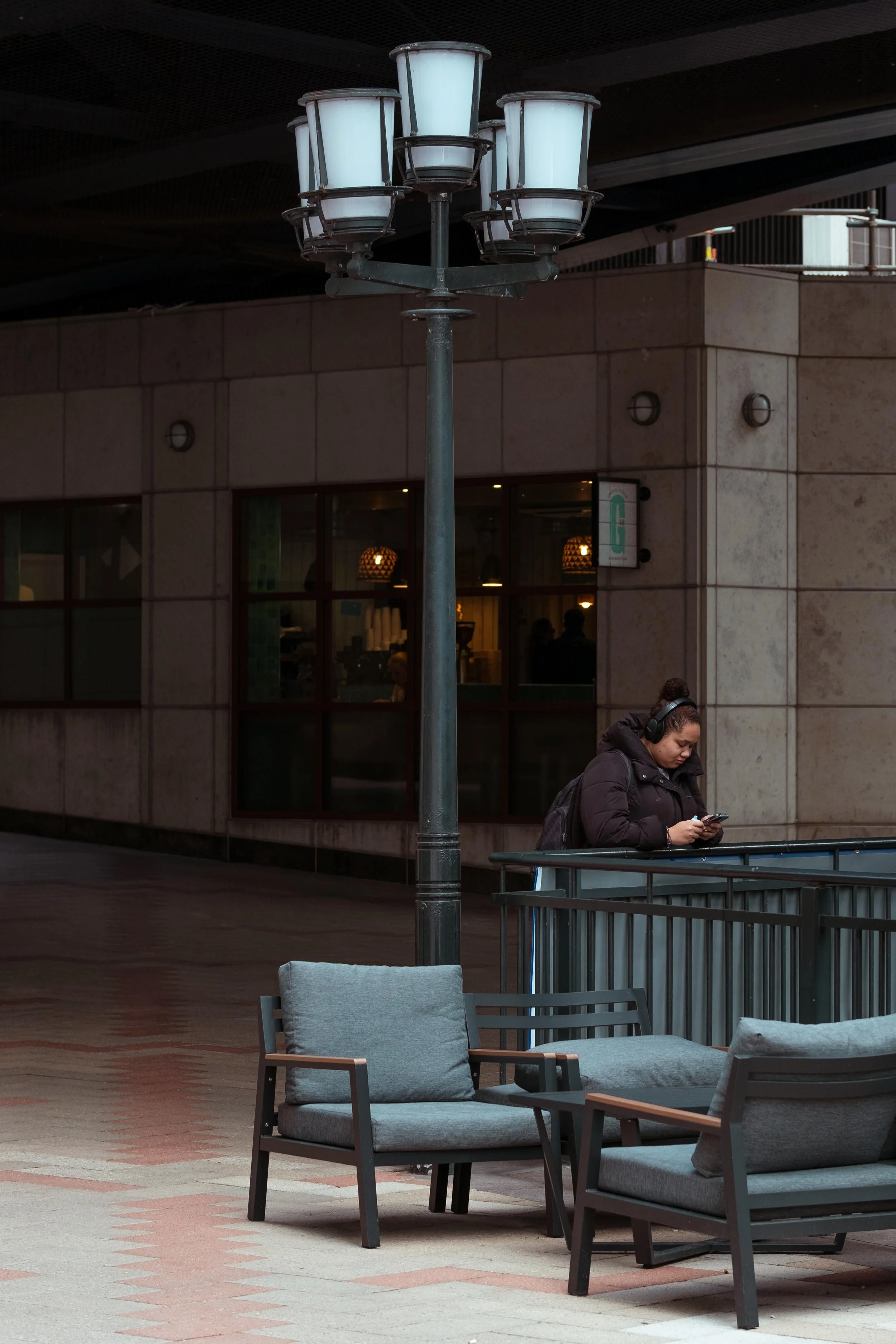 A woman with headphones standing outdoors, looking at her phone, near some outdoor chairs and a street lamp.