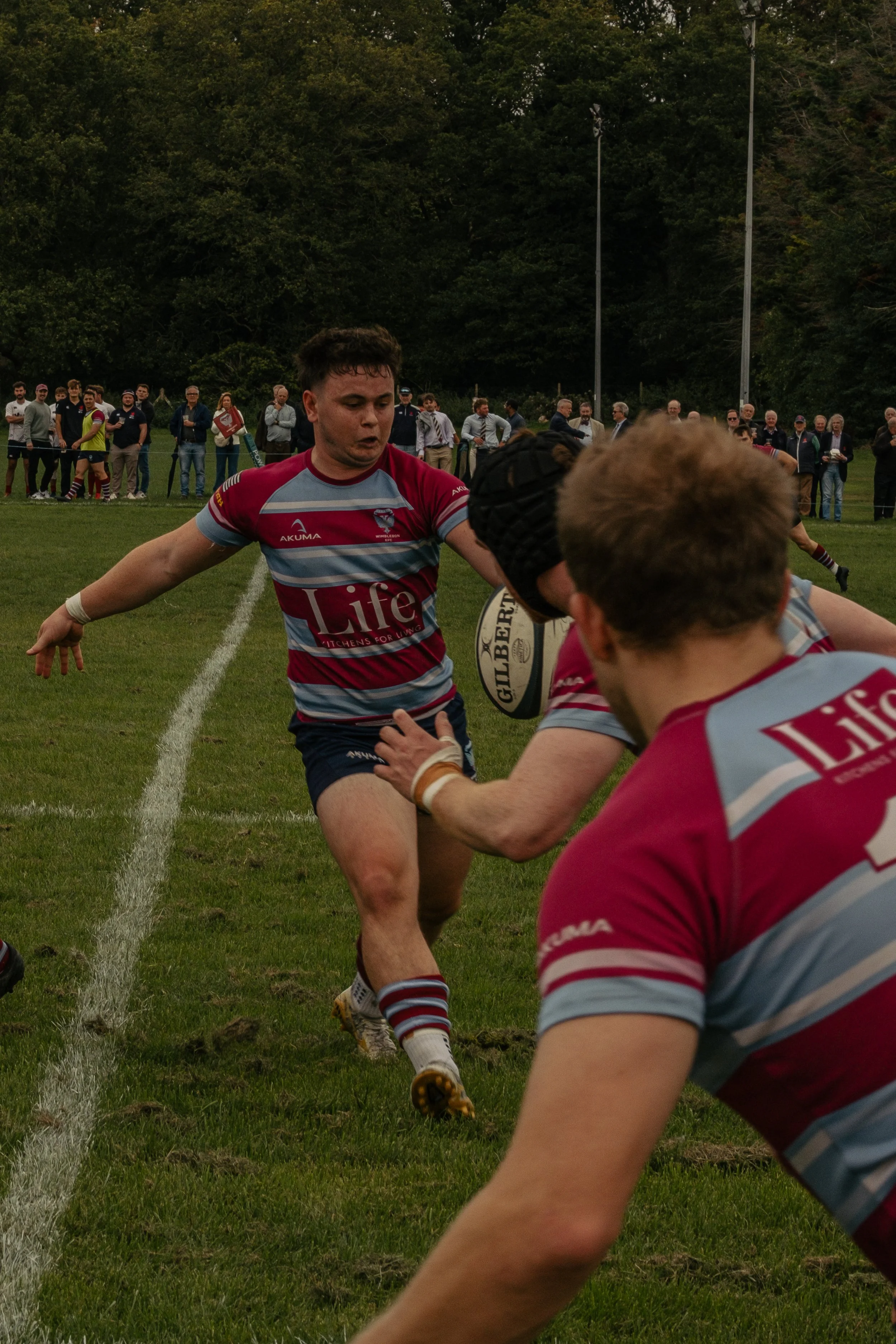 A rugby player in a maroon and gray striped uniform holding a rugby ball during a game, with other players and spectators watching on a grassy field.