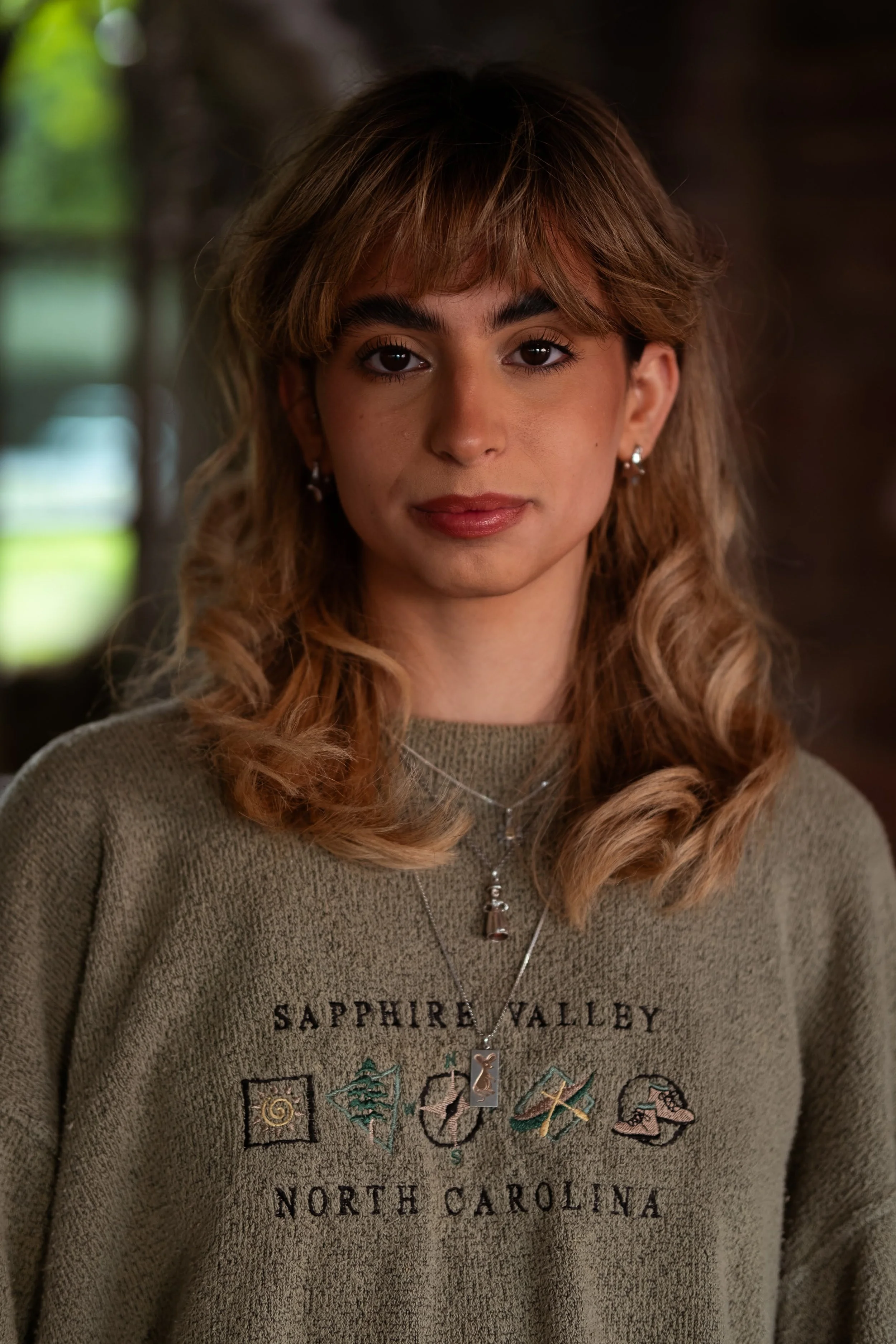 A young woman with curly, shoulder-length hair and earrings, wearing a greenish sweatshirt with 'Sapphire Valley North Carolina' embroidered on it, and layered necklaces, standing indoors with a blurred window and greenery in the background.