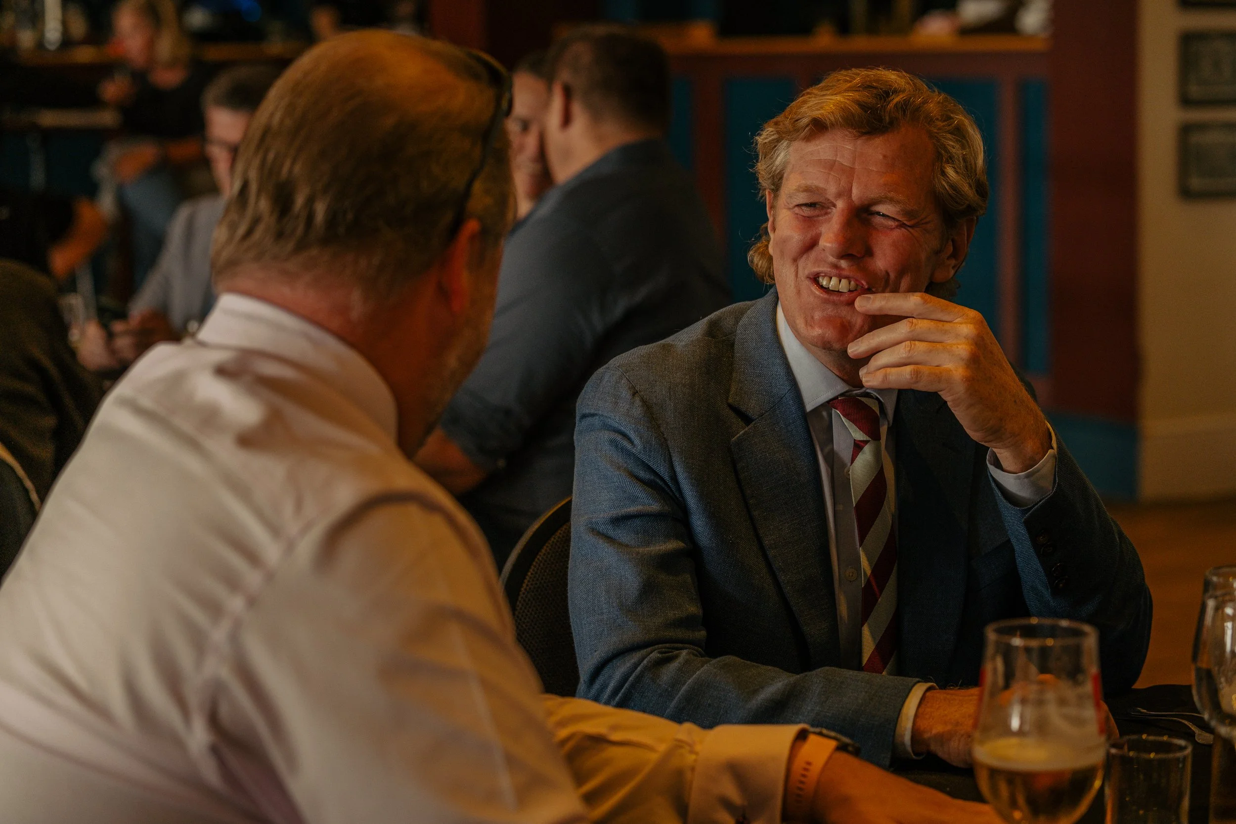 Two men in a restaurant or bar, engaged in a conversation. One is in a beige shirt, and the other in a dark suit with a striped tie, smiling and touching his face.