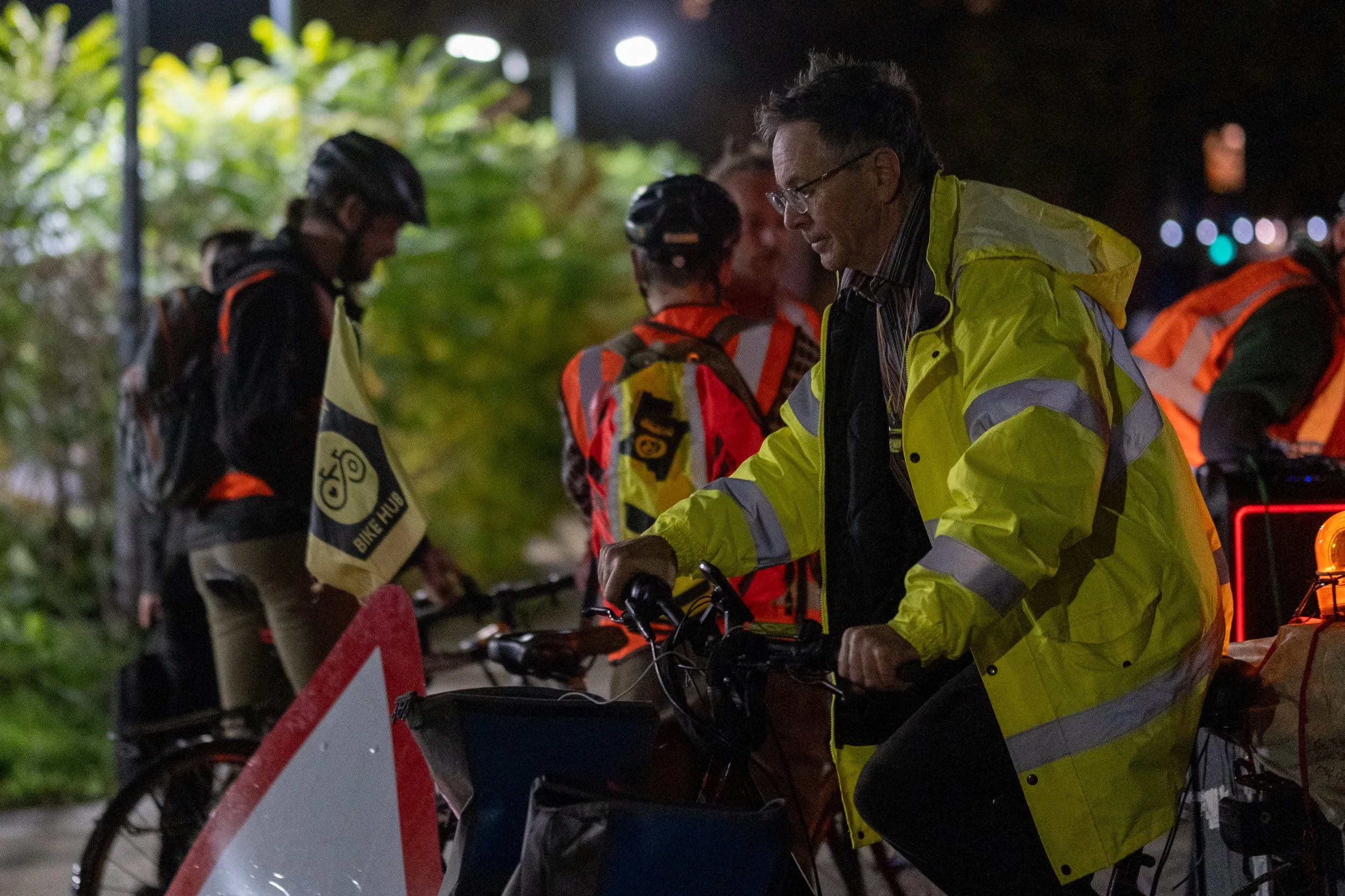 Nighttime scene of a group of cyclists, with one person in a yellow reflective jacket in the foreground, preparing or adjusting their bicycle, while others in orange and black reflective gear stand nearby.