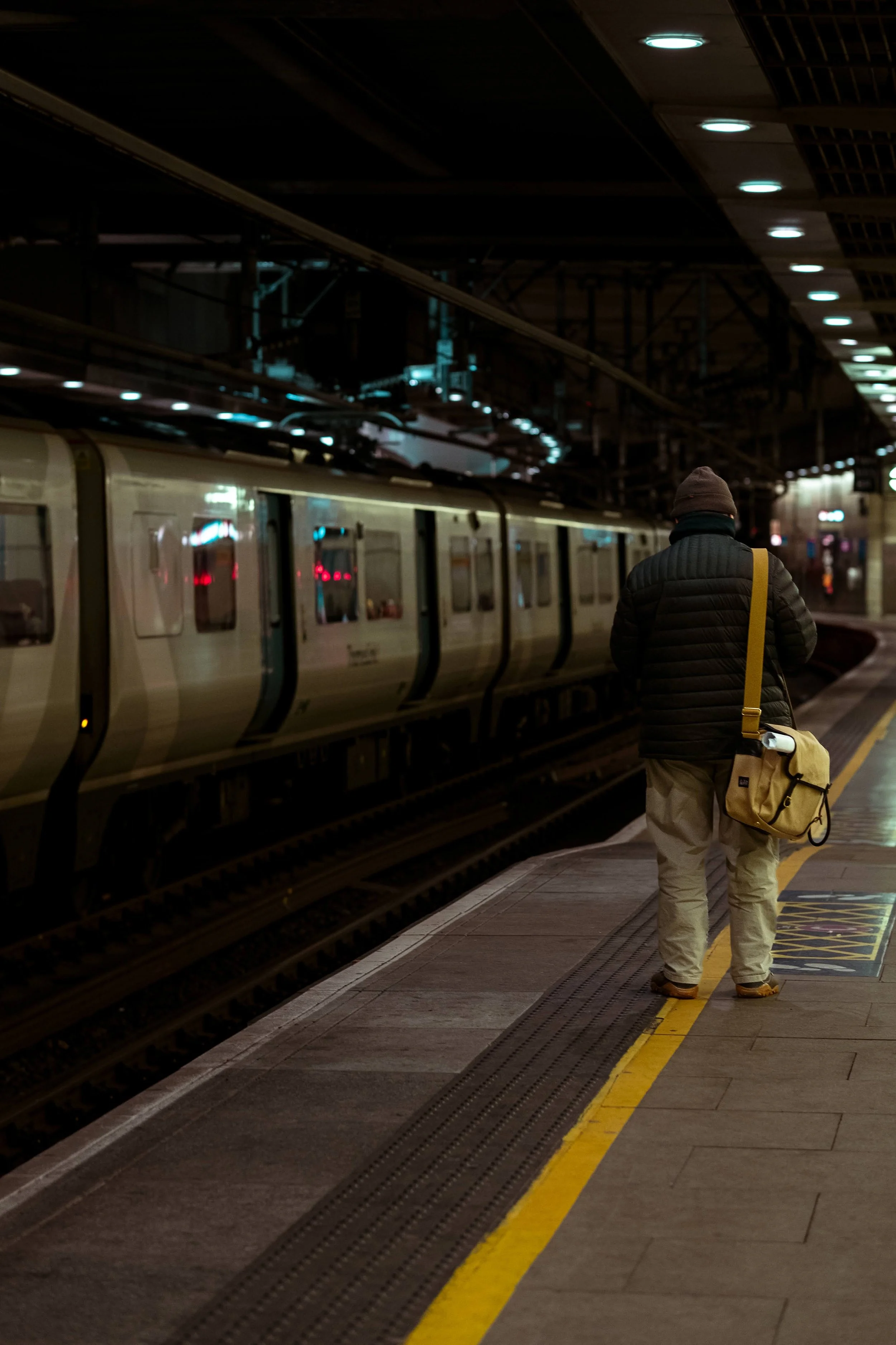 Person standing on an underground train platform, waiting for a train at night, with a beige handbag over their shoulder.