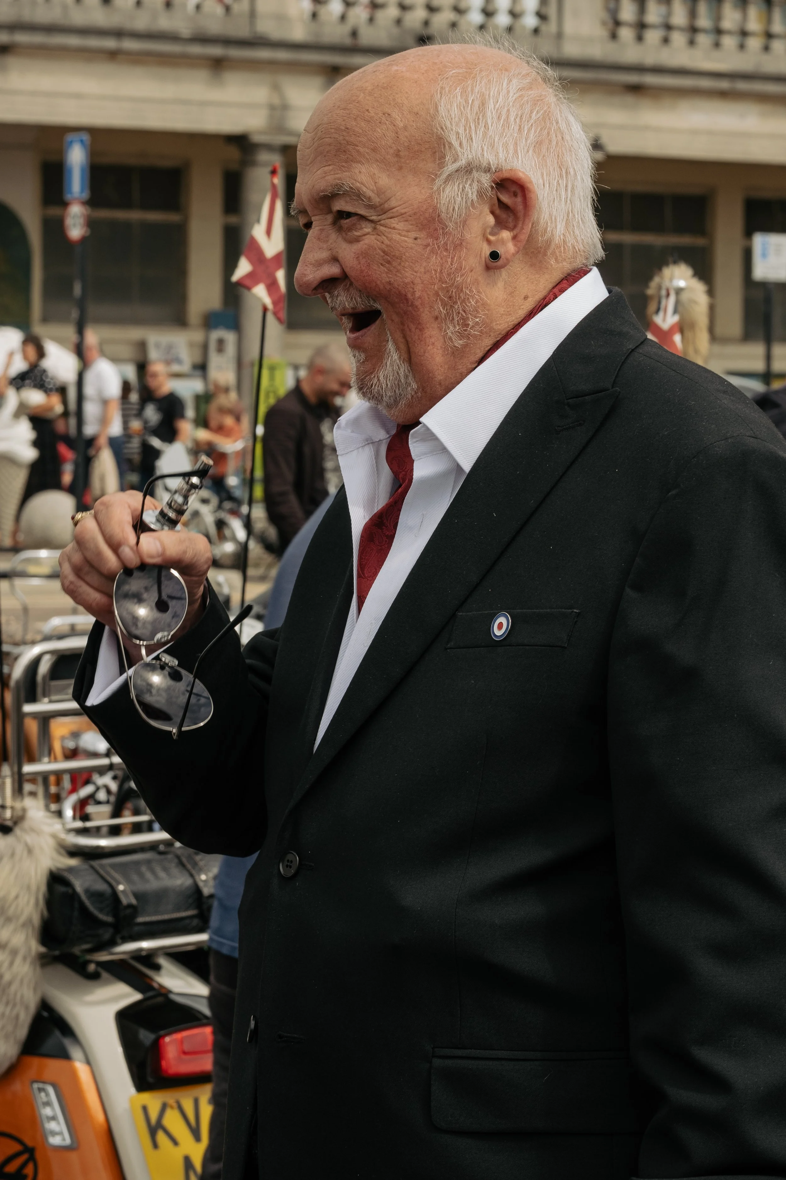 A smiling elderly man with white hair and beard, wearing a black suit, white shirt, and red tie, holding sunglasses in his right hand at an outdoor event.