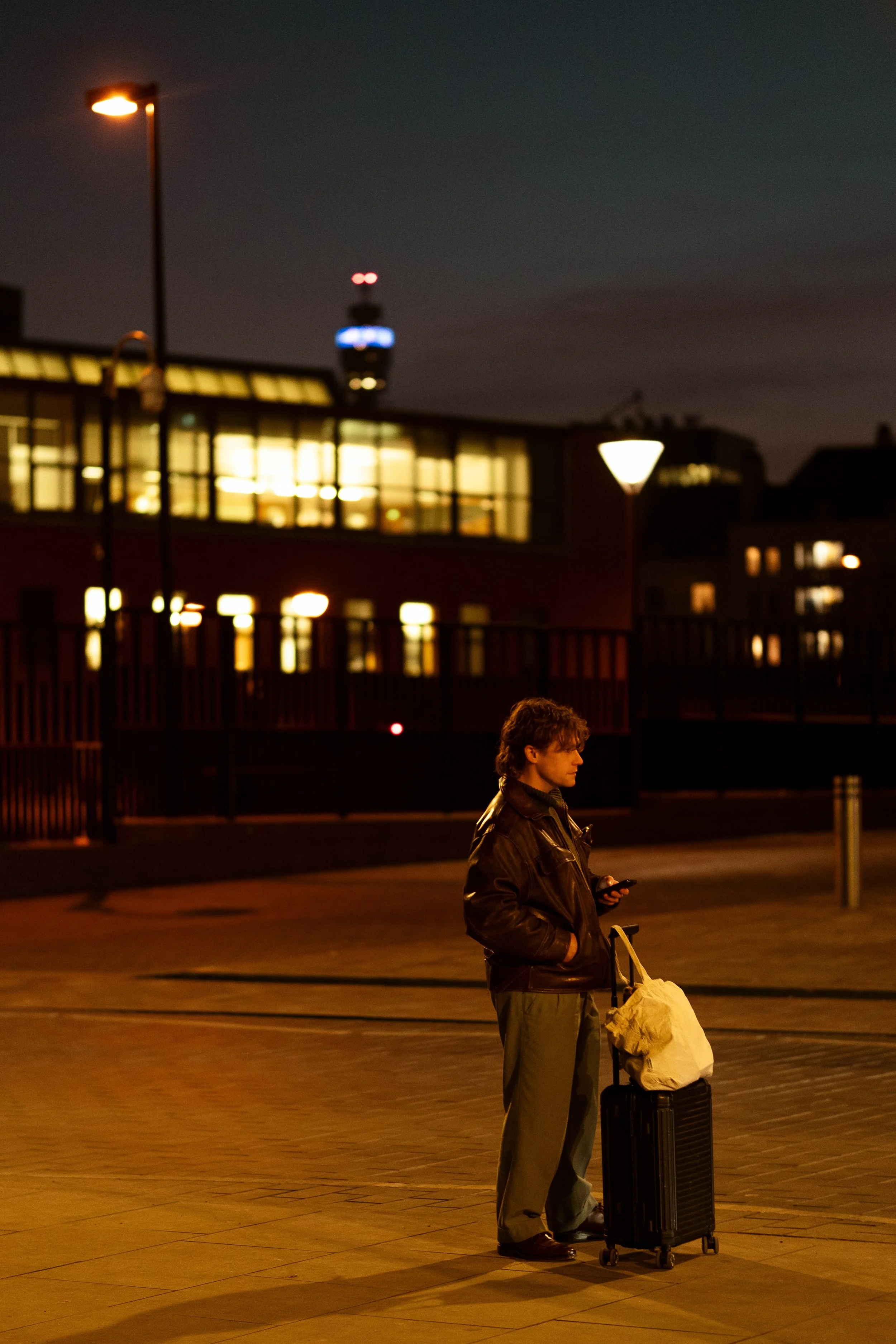 A man standing alone on a city street at night with a rolling suitcase, looking at his phone.