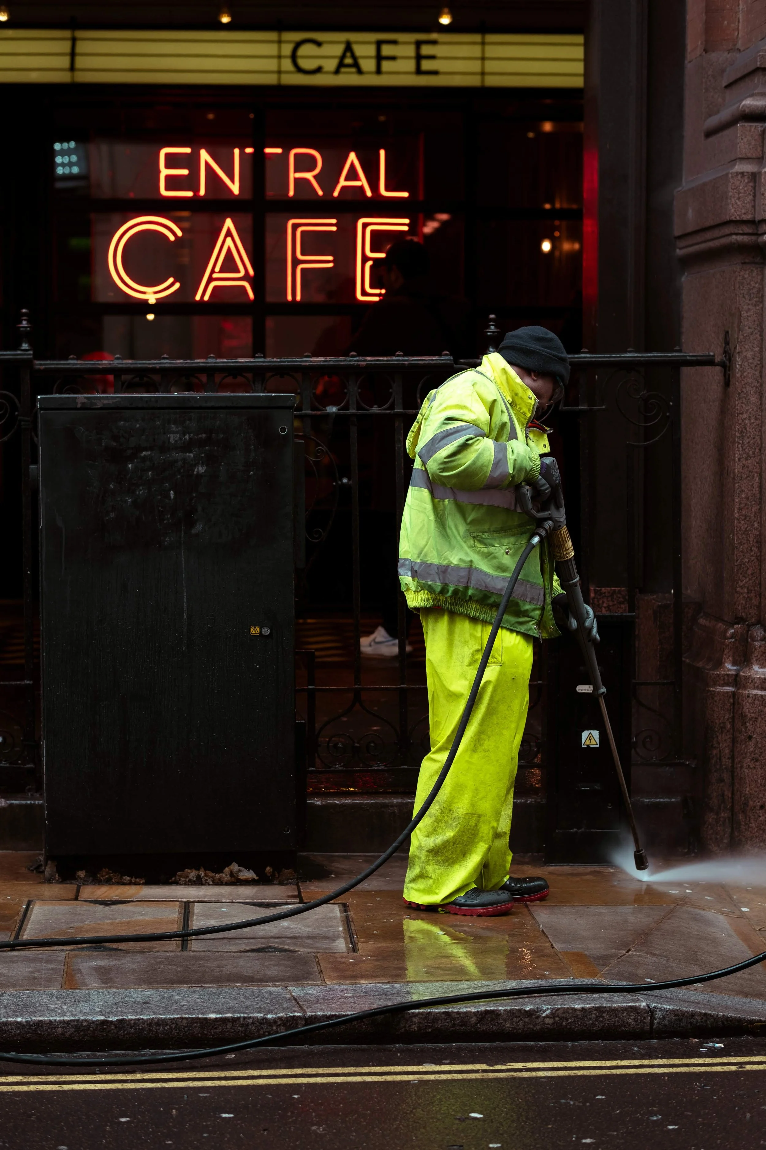 A worker in yellow high-visibility clothing and black gloves pressure washing the sidewalk outside a cafe with a yellow sign and illuminated red sign that says "Central Cafe."