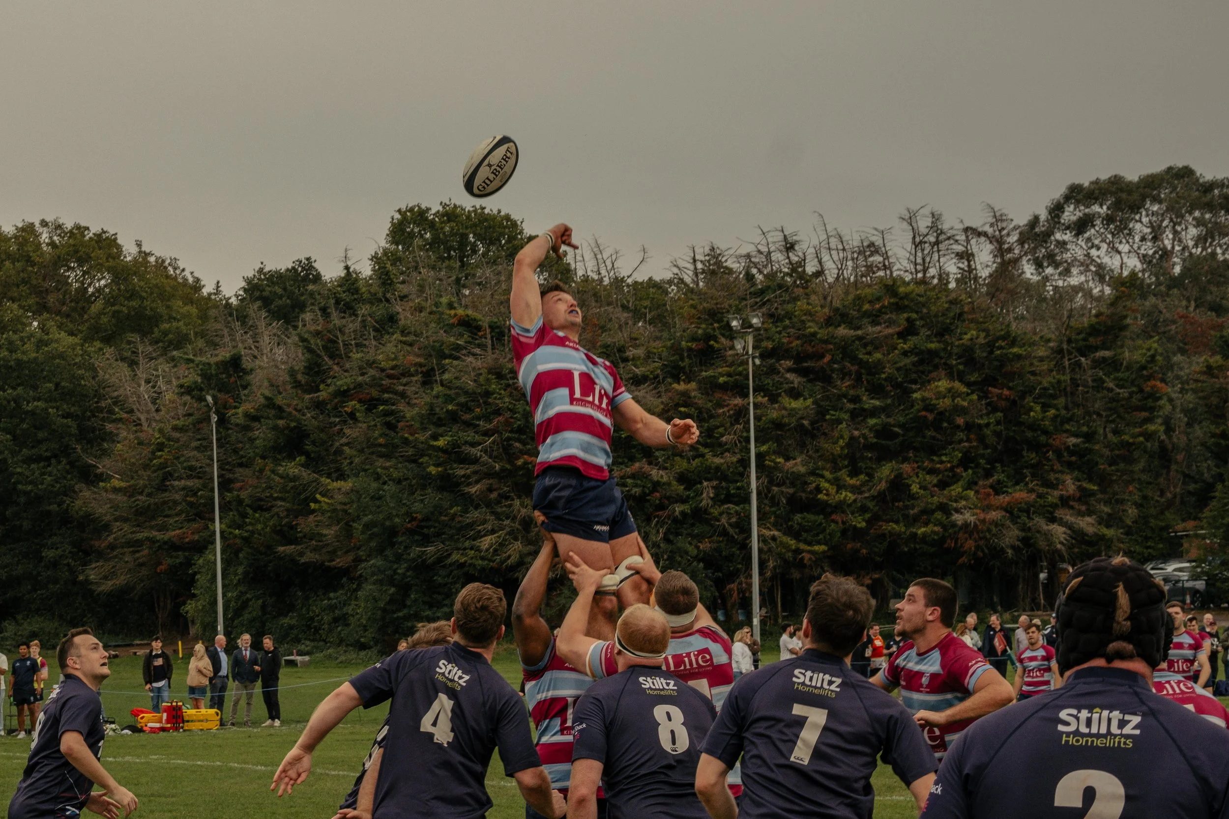 Rugby players in a lineout competition, with one player being lifted to catch the ball, during a game on a grassy field with a background of trees and spectators.