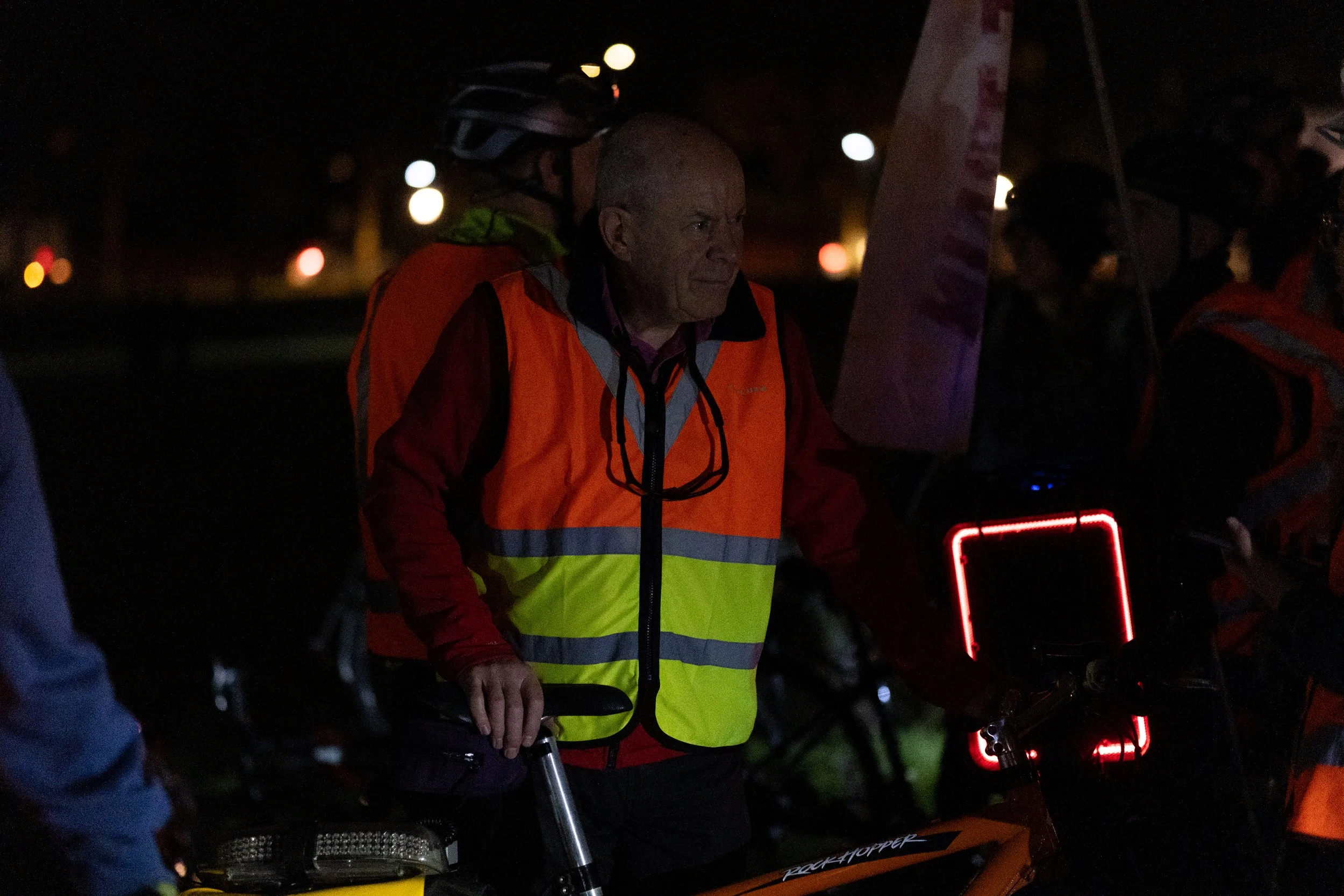 Man wearing a reflective safety vest standing next to a bicycle at night, with other people around him also in safety gear.