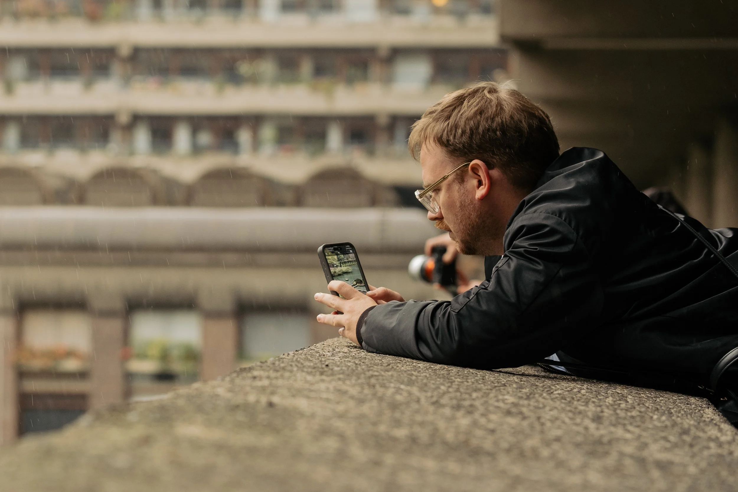 A man with glasses and a black jacket lying on a concrete surface using a smartphone, with a multistory building in the background.