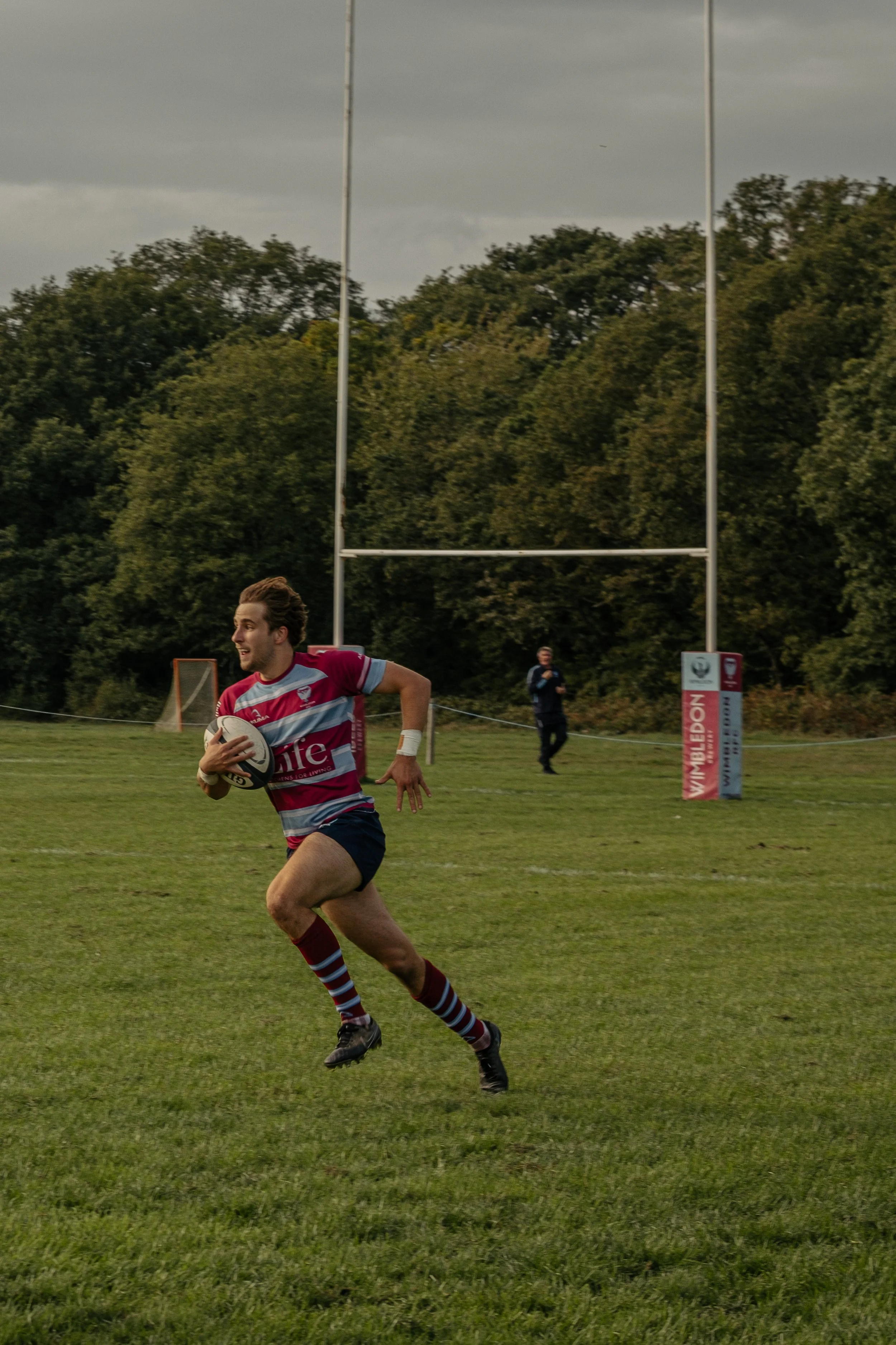 A rugby player running on a field holding a rugby ball, with goalposts and an official or coach in the background.