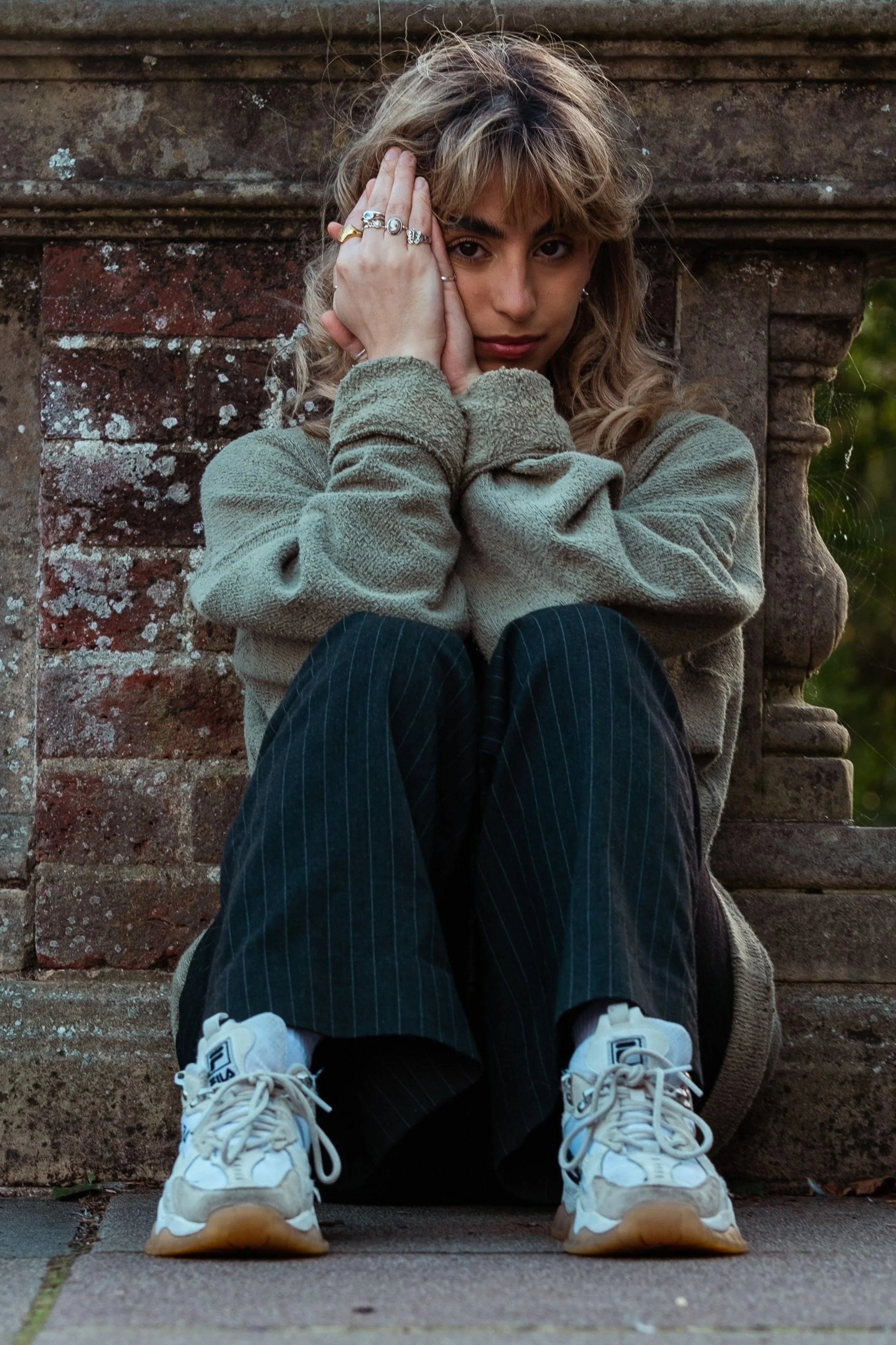A young woman with curly blonde hair sits on the ground, leaning against a brick and stone wall, with her knees drawn up and hands framing her face, wearing a beige fleece jacket, black pinstripe pants, and white sneakers.