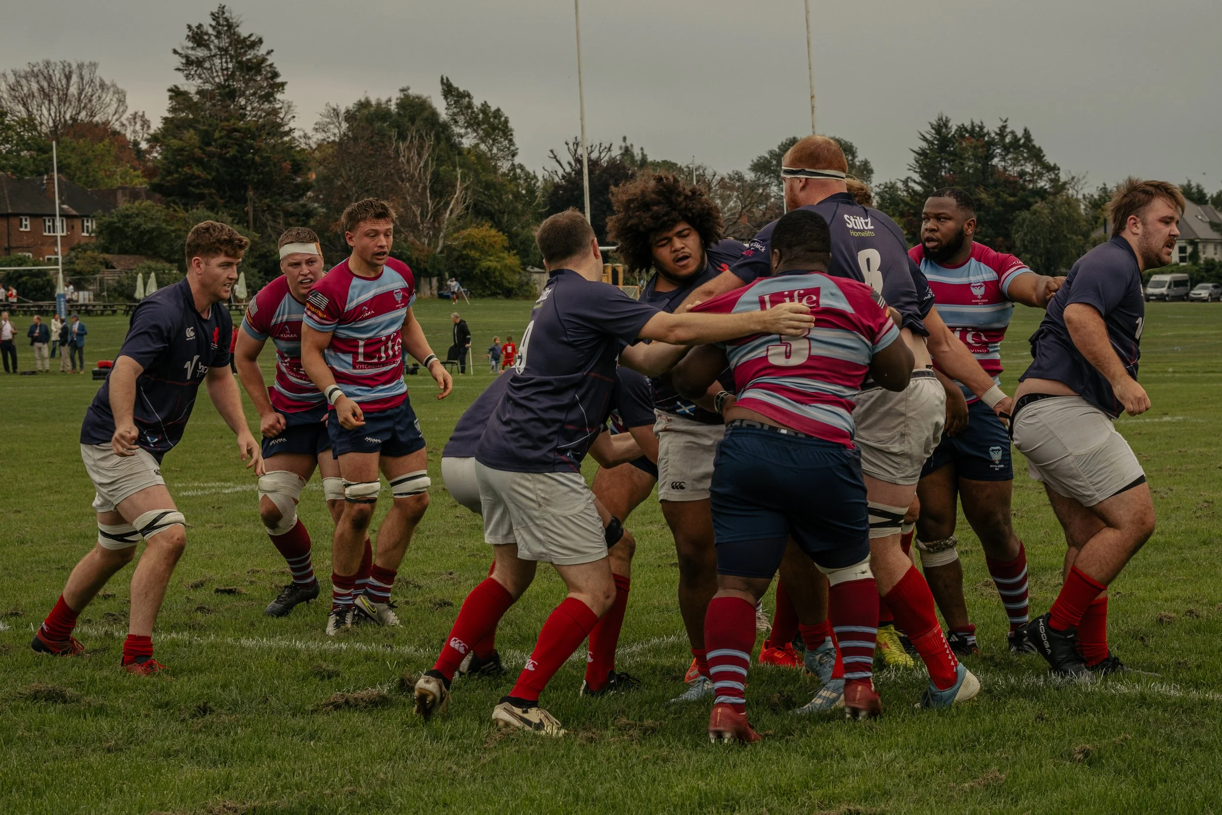 Rugby players in a scrum during a match on a grassy field with spectators and houses in the background.