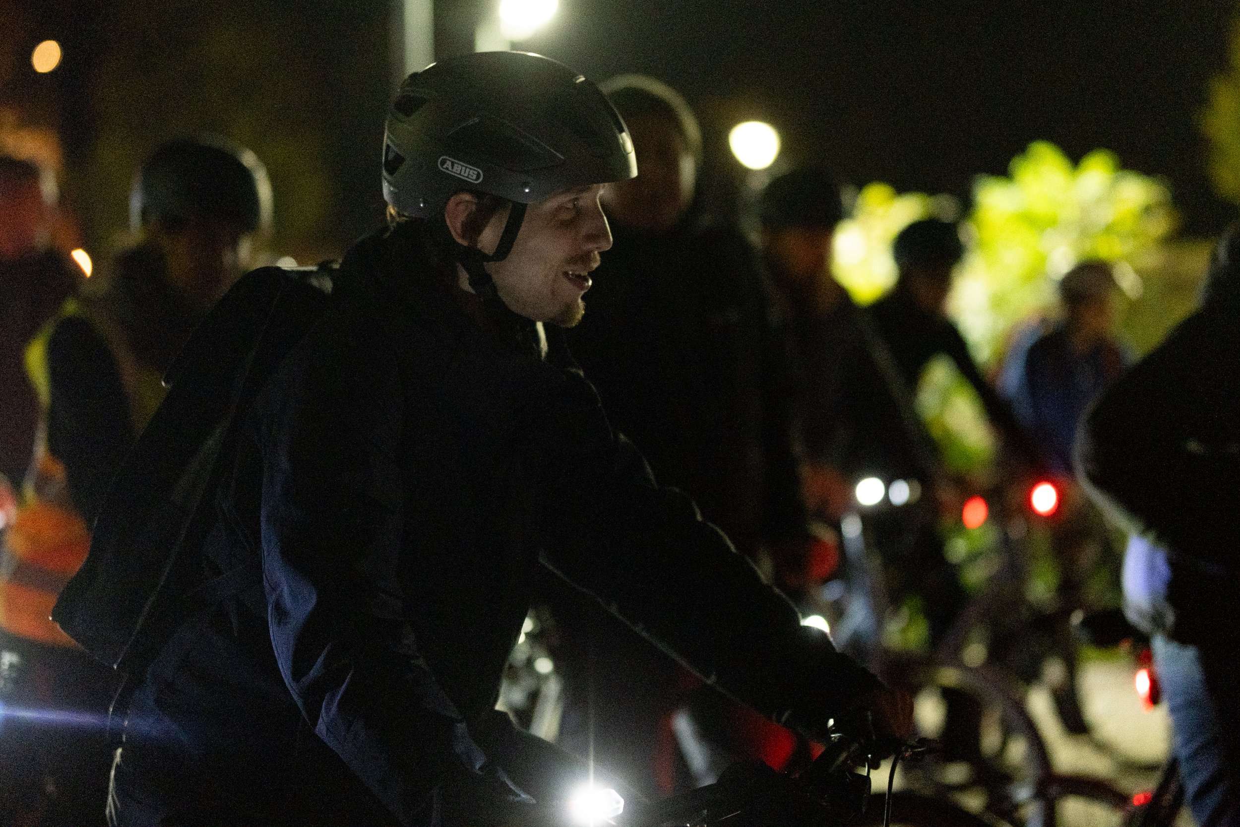 A man with a helmet riding a bicycle at night, with a group of people and blurred lights in the background.