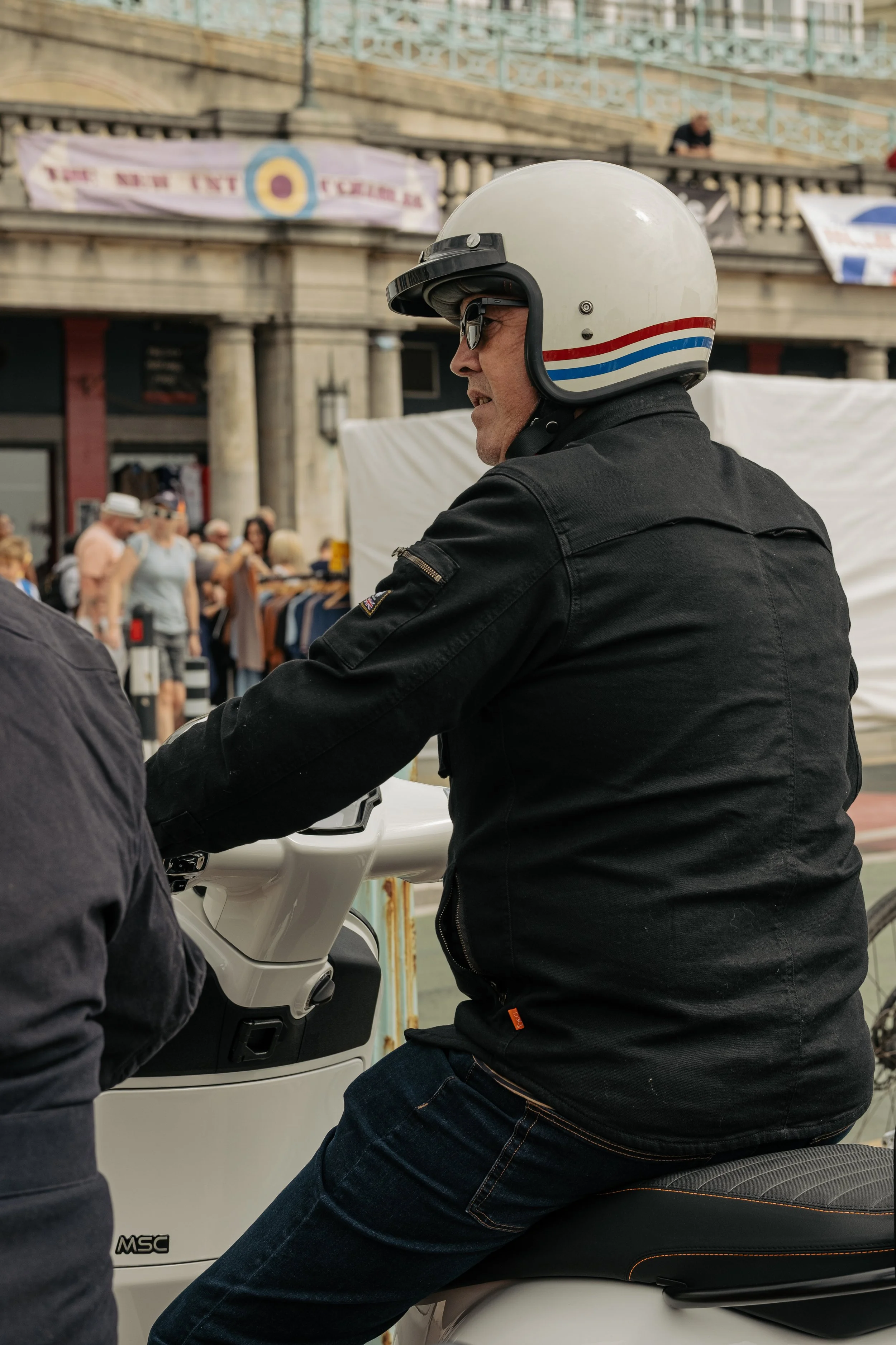 A man wearing a white helmet and black jacket is sitting on a white scooter in a crowded outdoor area with people and a building in the background.