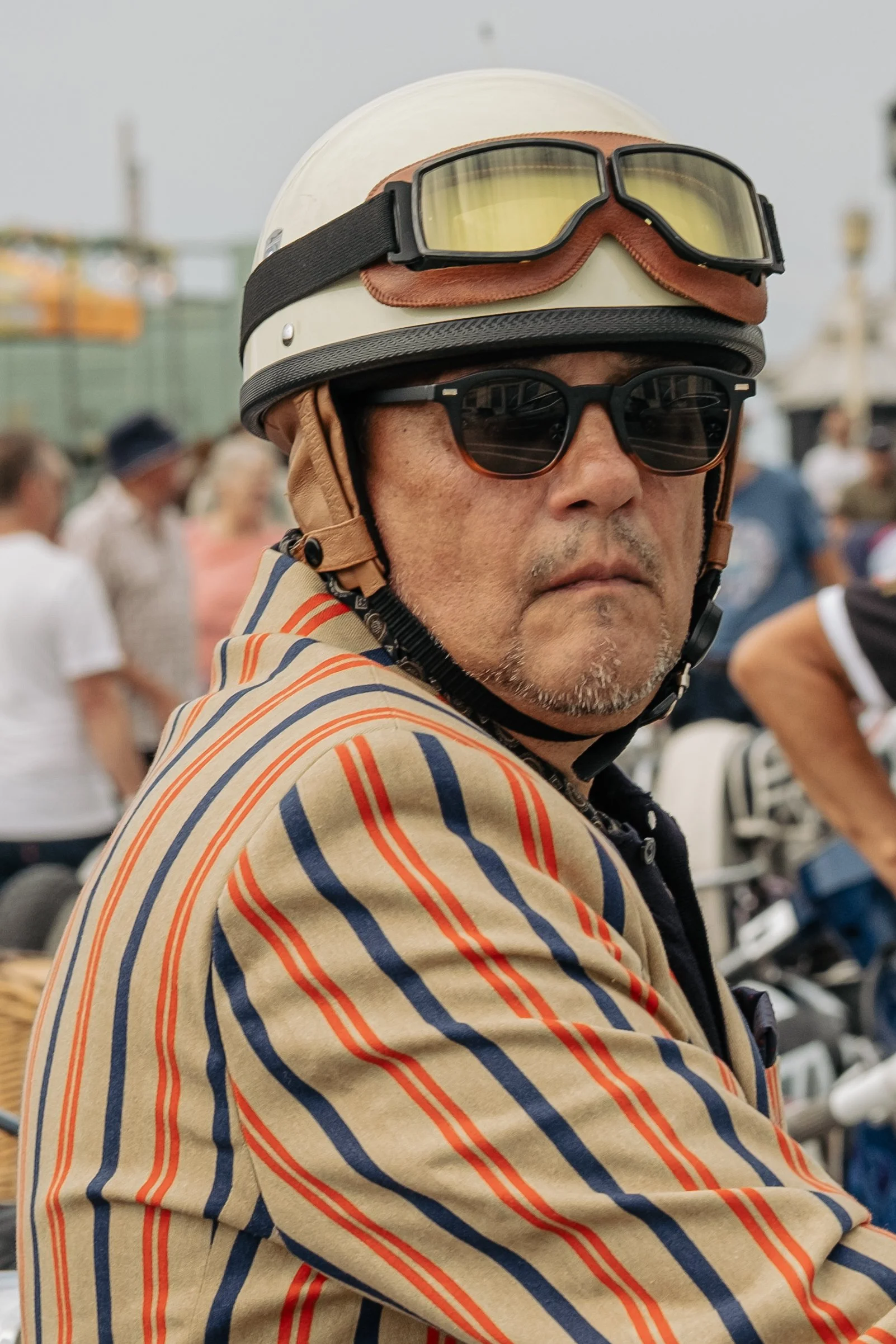 Close-up of a man wearing sunglasses, a vintage-style helmet with goggles on top, and a colorful striped shirt, at an outdoor event with other people in the background.
