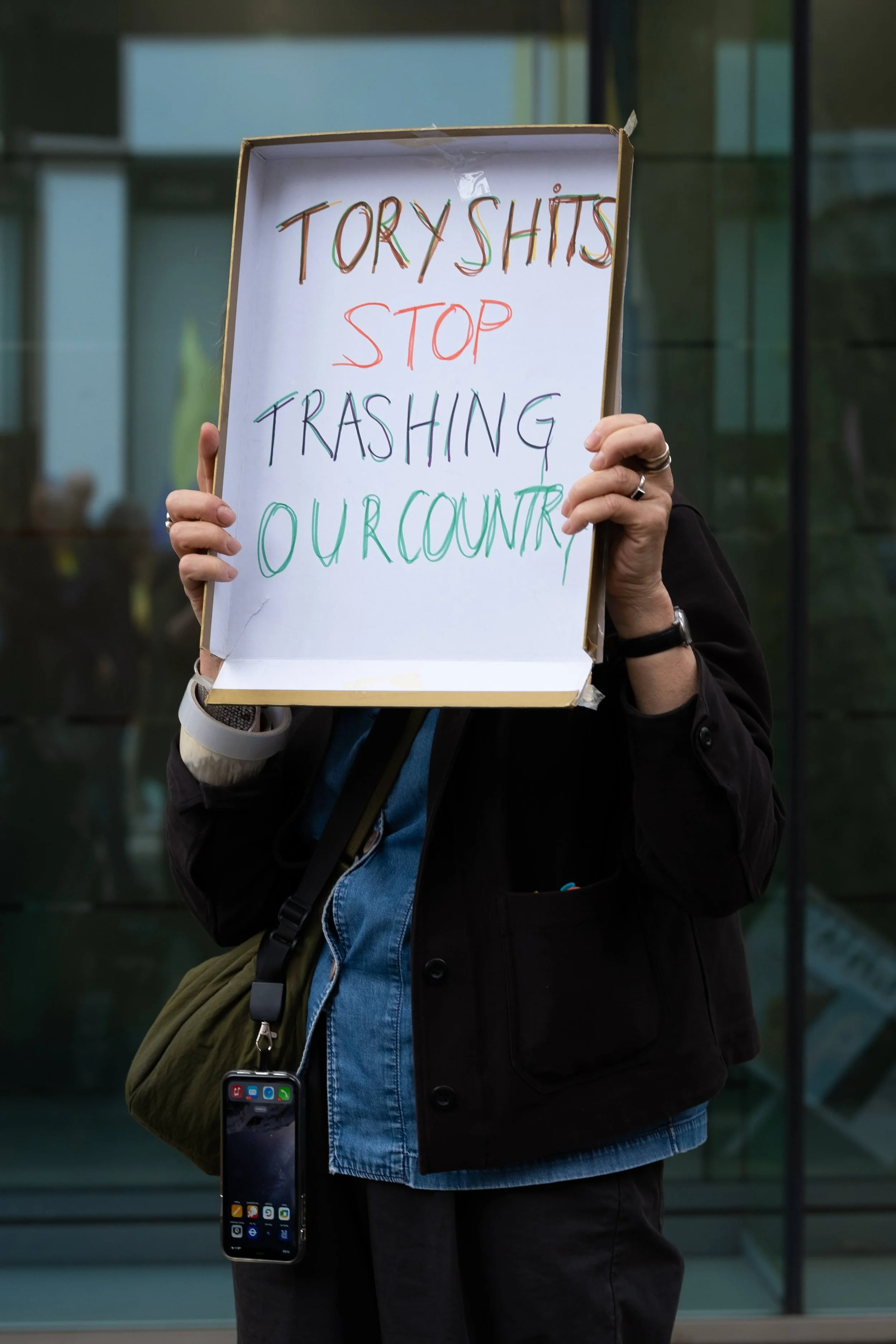 Person holding a protest sign that reads, "TORY SHITS STOP TRASHING OUR COUNTRY."
