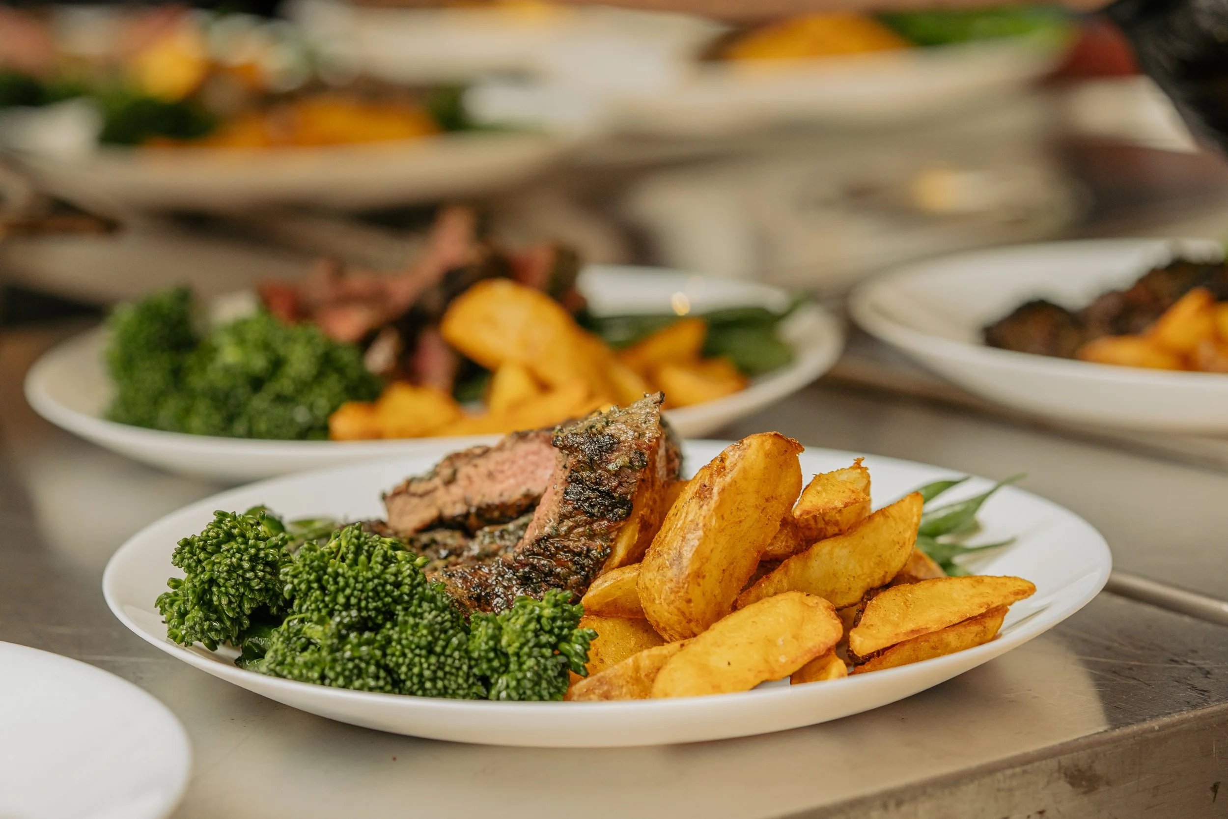 Plate with seasoned potato wedges, grilled steak, and broccoli, with more similar plates in the background.