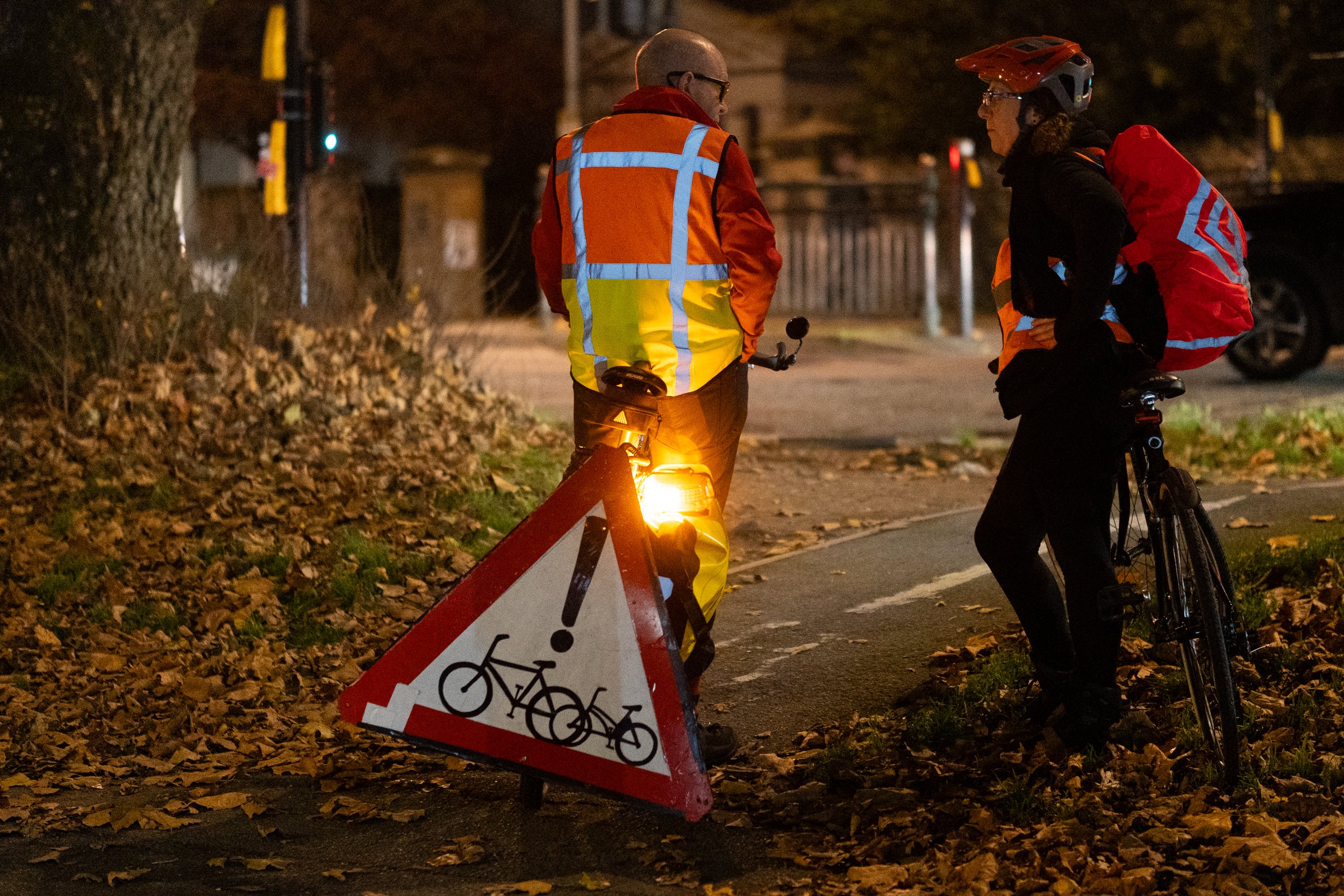 Two cyclists talking on a bike path at night, one with a reflective vest and the other with a helmet and backpack, near a warning sign for bicycles.