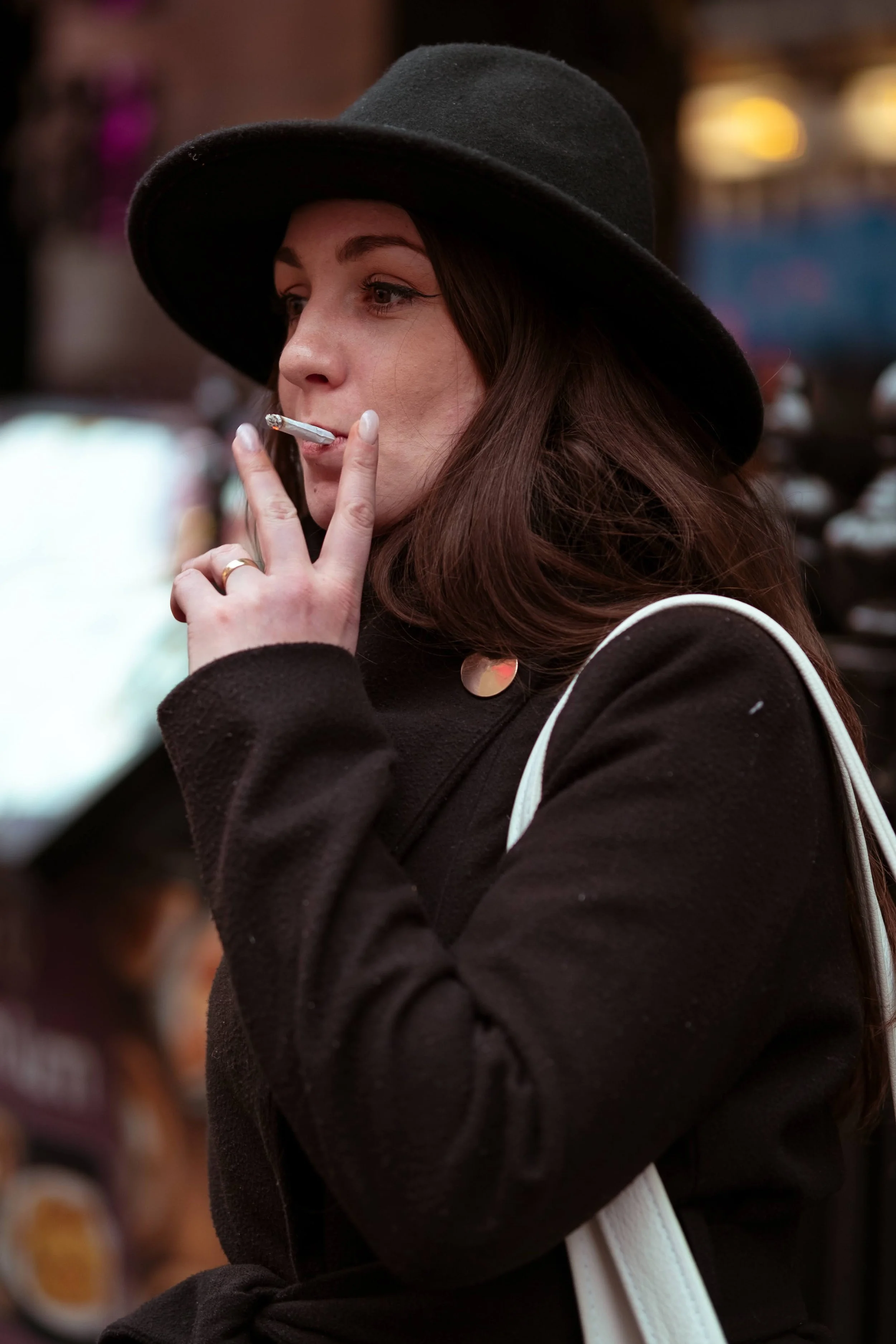 A woman with long brown hair wearing a black hat and black coat, smoking a cigarette, in an urban setting.