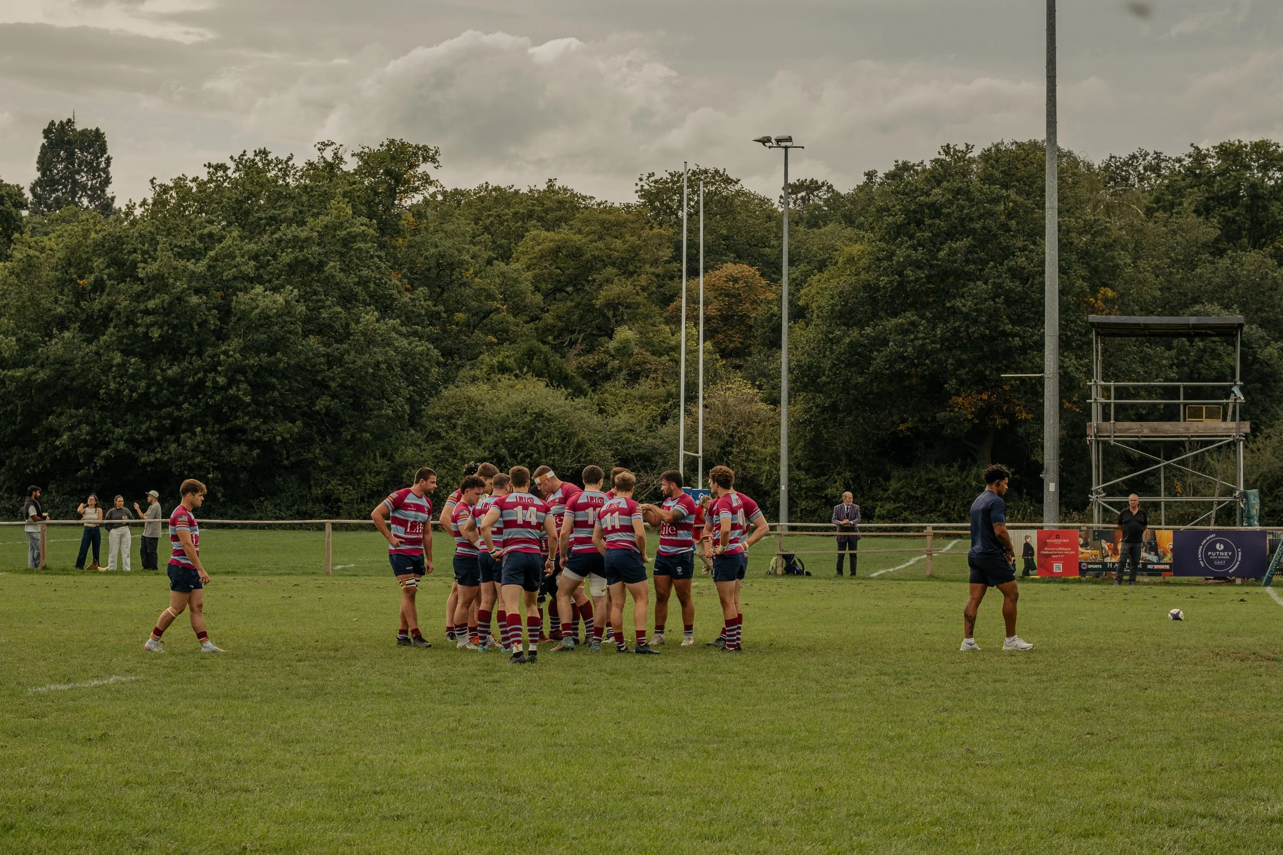 Rugby team in striped jerseys huddled together on a grassy field during a game, with few spectators and trees in the background.