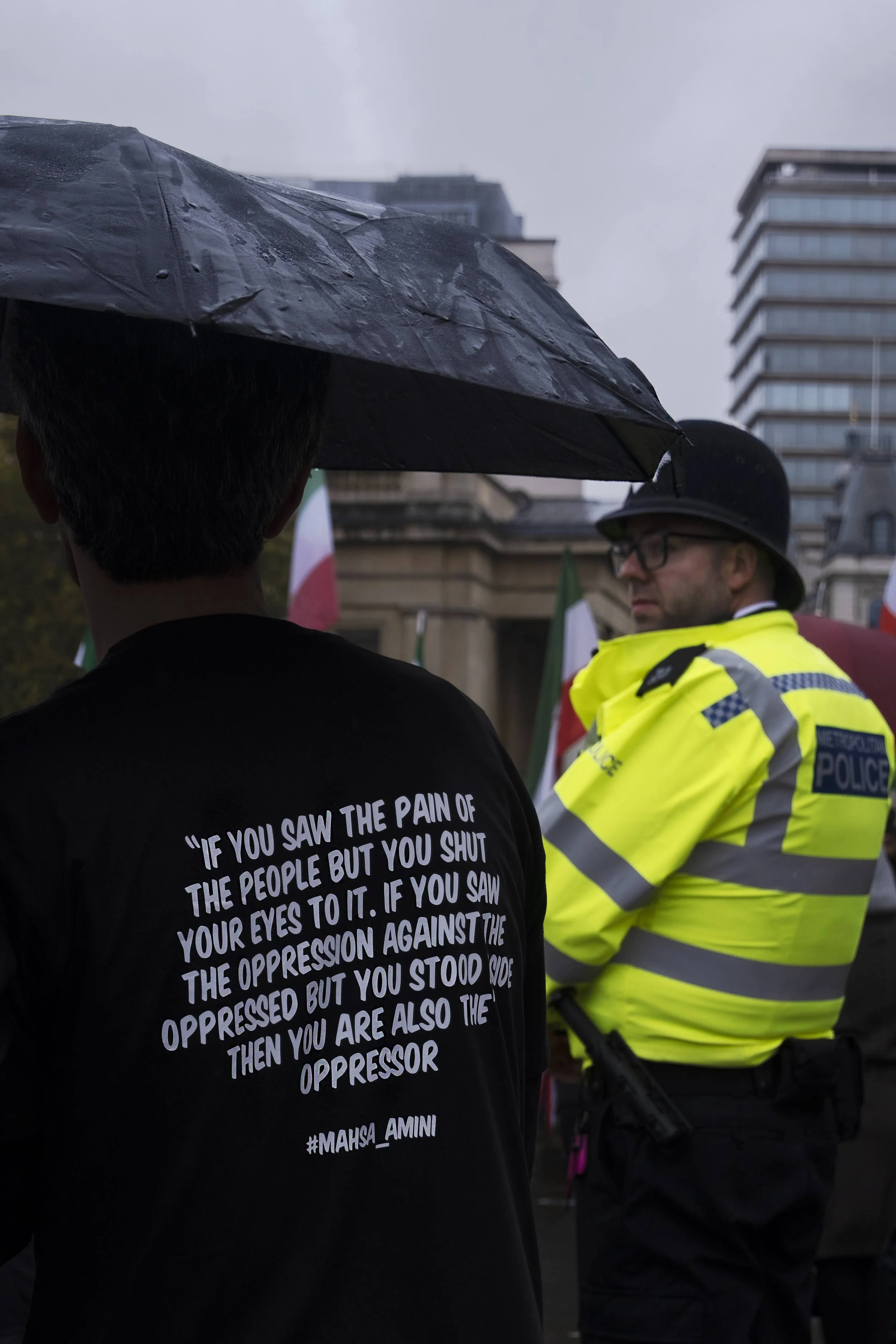 A person with a black shirt bearing a quote standing near a police officer during a rainy protest, with flags and a building in the background.