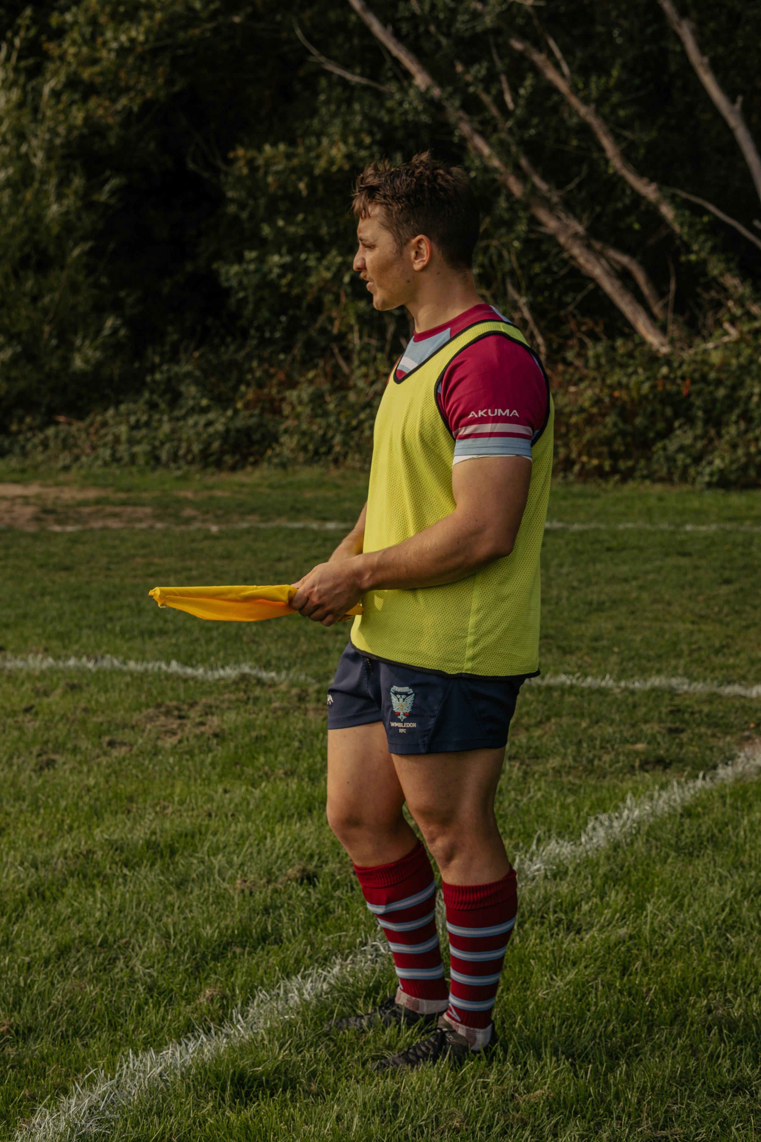 A rugby player standing on a grassy field, holding a yellow flag, dressed in a yellow training vest, maroon and grey striped socks, black soccer cleats, and navy blue shorts with a team logo.