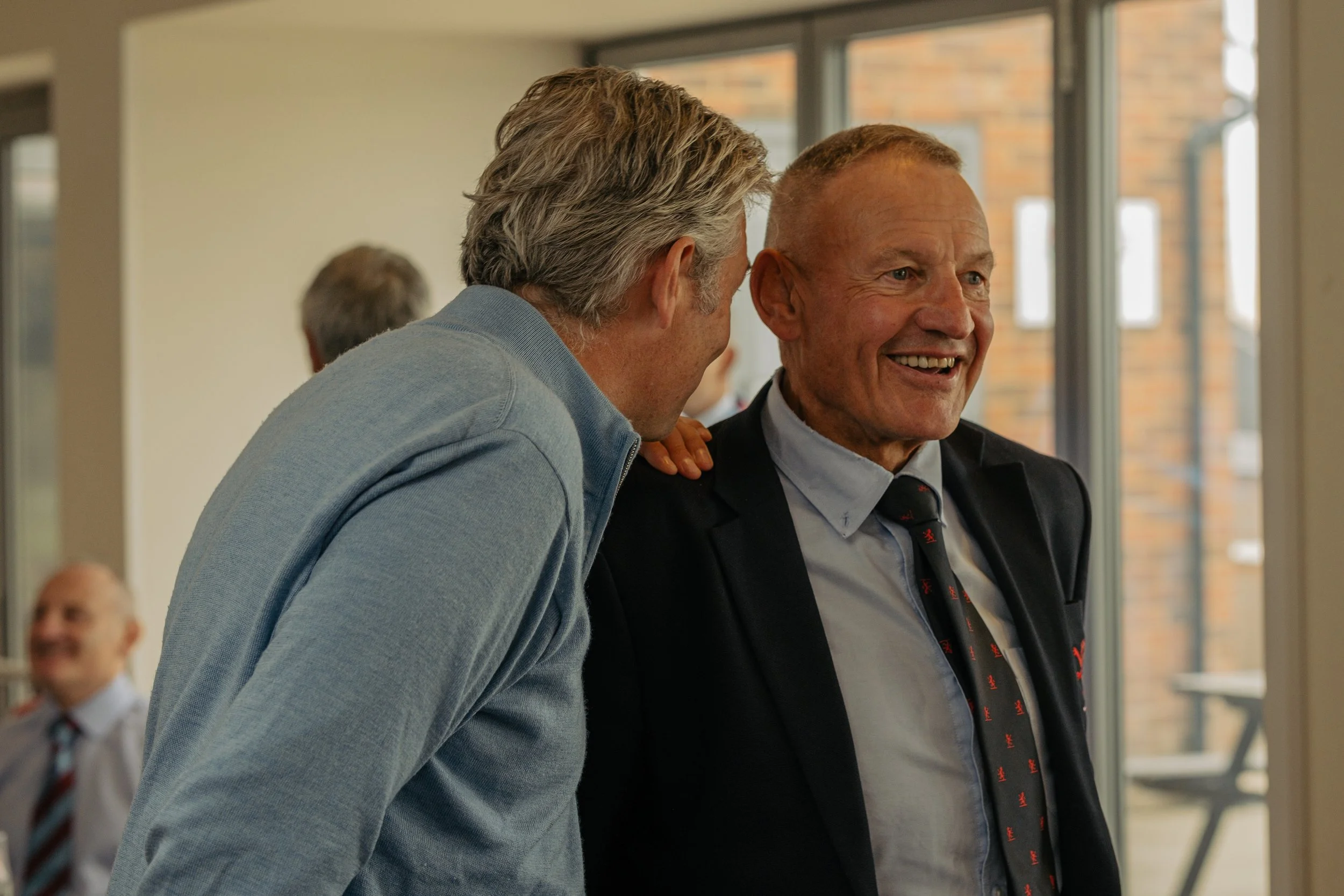Two middle-aged men in business attire smiling and talking at a social gathering indoors, with other people in the background.