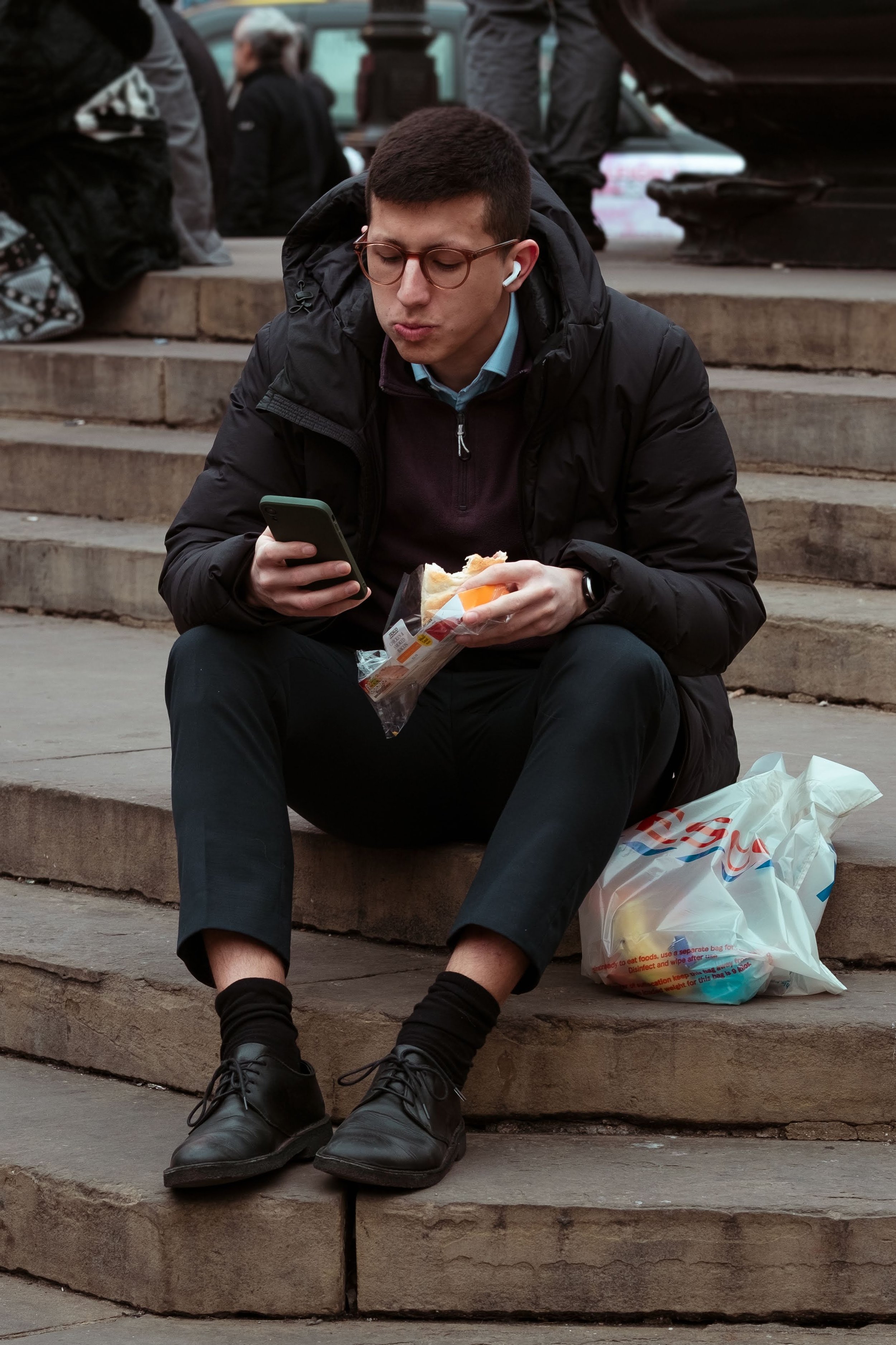 A young man sitting on outdoor stone steps, wearing glasses, a black jacket, black pants, and black shoes. He is holding a sandwich in one hand and a smartphone in the other, with earbuds in his ears. A plastic grocery bag is next to him on the steps