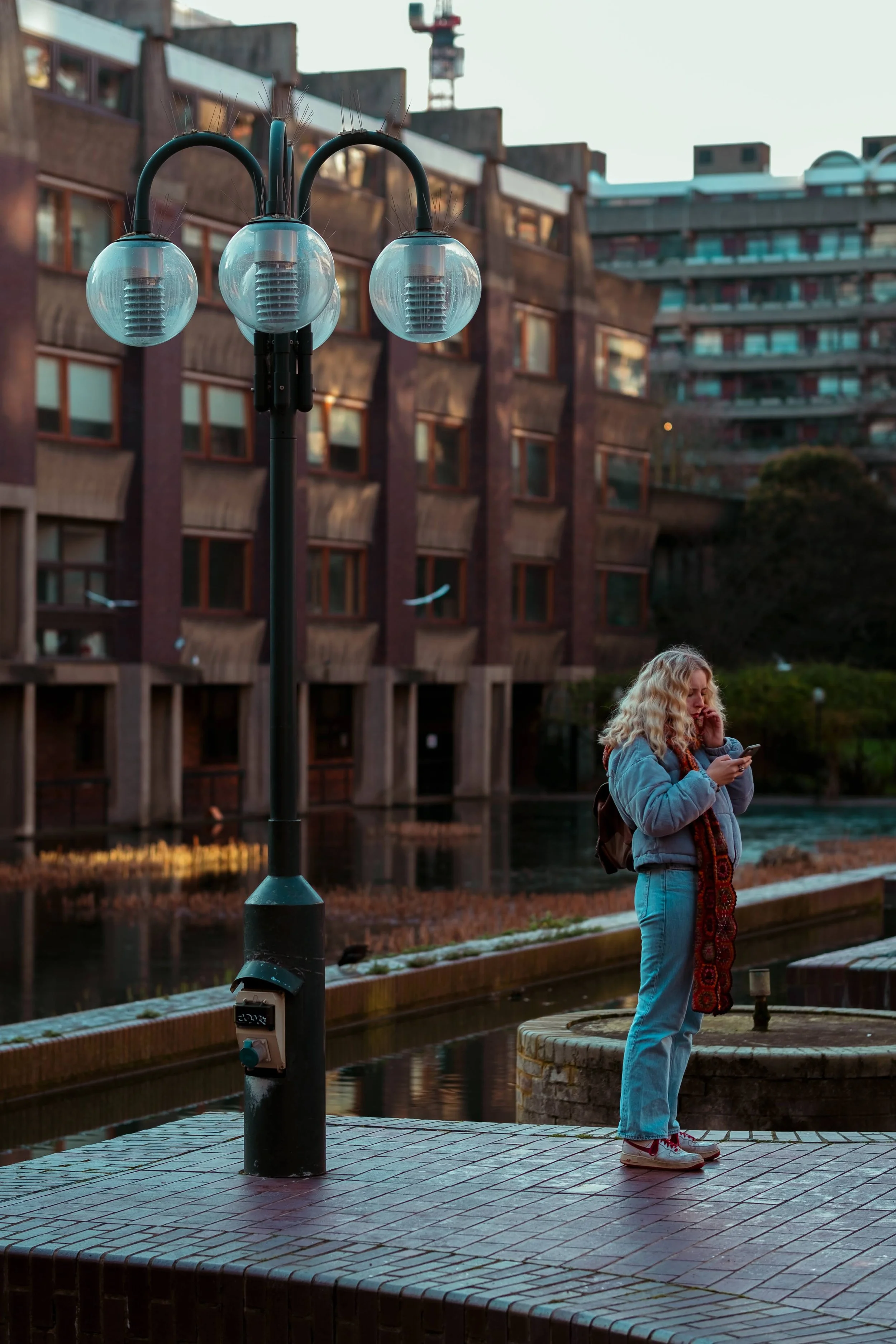 A woman with curly blonde hair wearing a blue jacket and jeans, standing on a paved walkway near a body of water, looking at her phone. There is a decorative street lamp with three round glass bulbs in the foreground and modern apartment buildings in