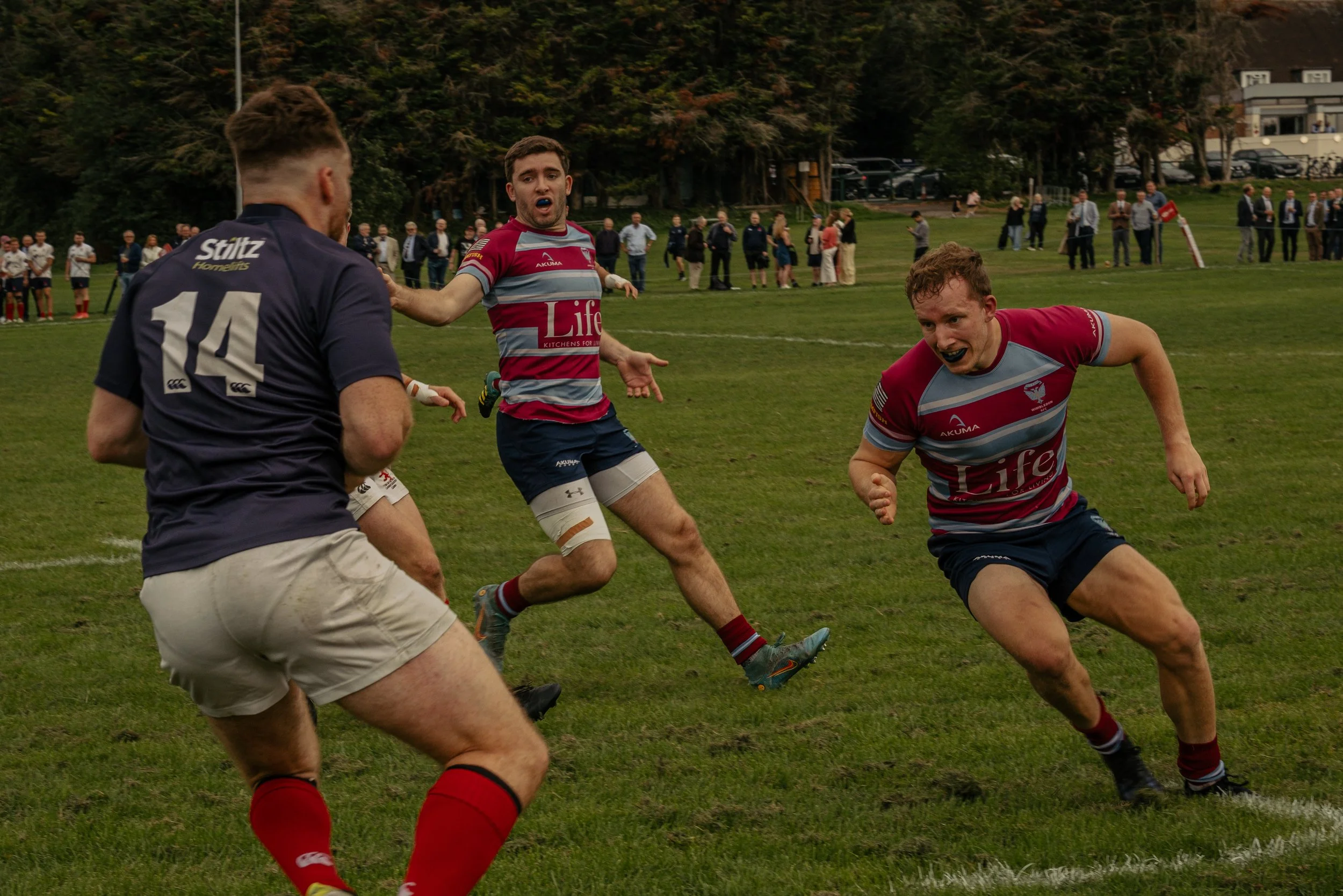 Rugby players in action on a grass field, with onlookers in the background.