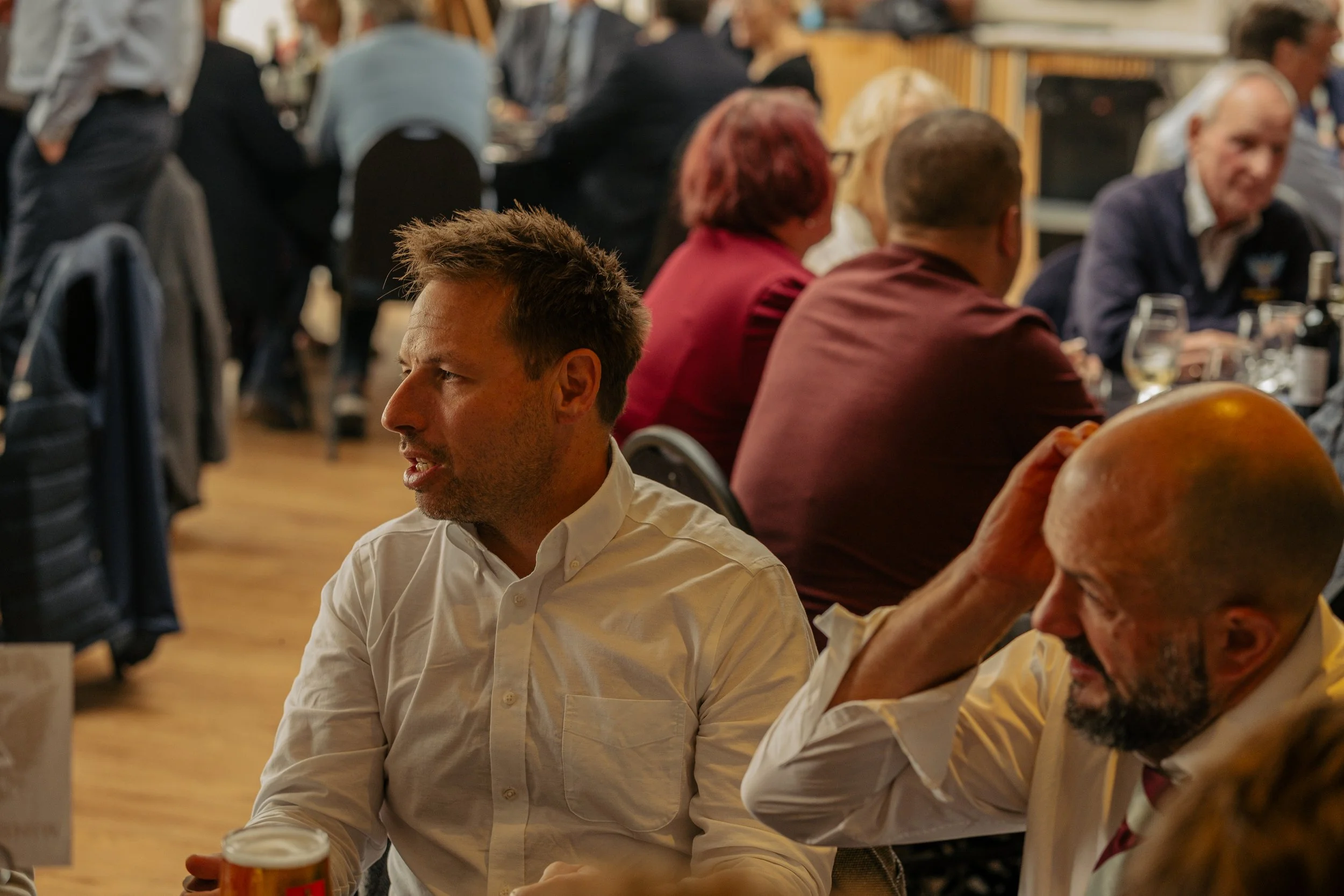 People sitting at tables in a busy restaurant or dining area, engaging in conversation, with glasses of wine and food on the tables.