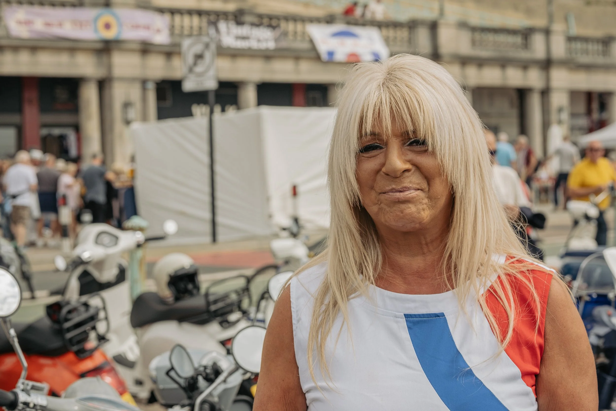 A woman with long blonde hair and a white, blue, and red sleeveless top, standing outdoors near parked motorcycles and a crowd of people in an urban setting.