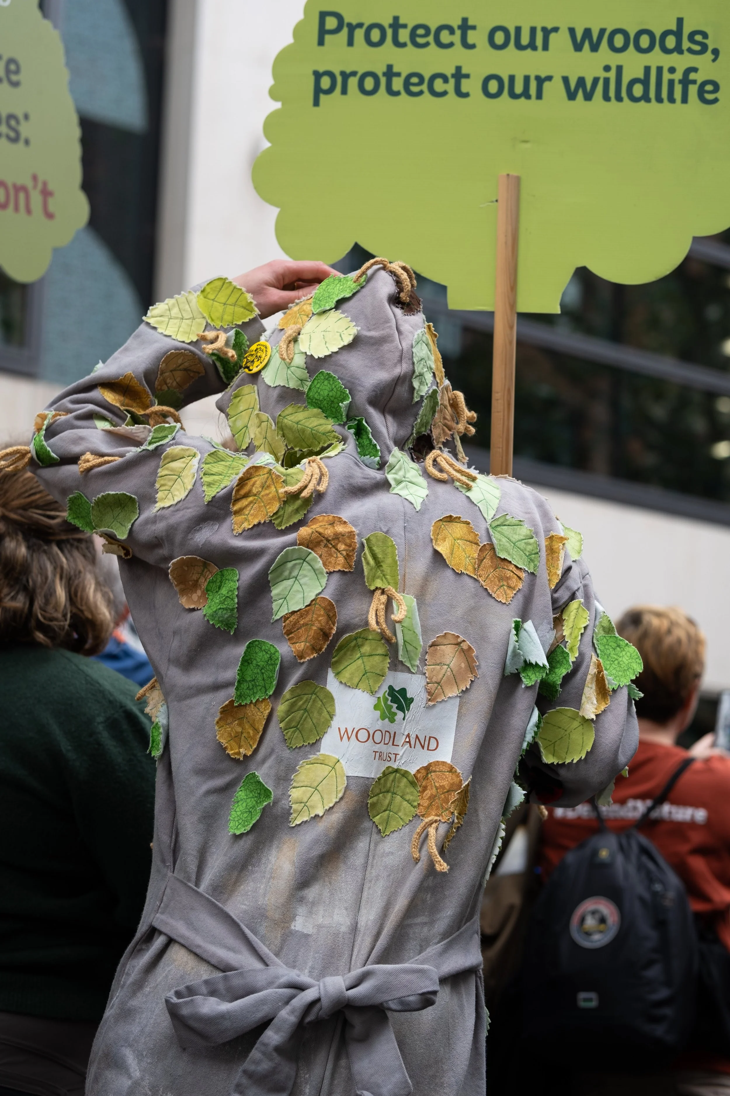 Person in costume covered with faux leaves at an environmental awareness event, holding a sign that reads "Protect our woods, protect our wildlife."
