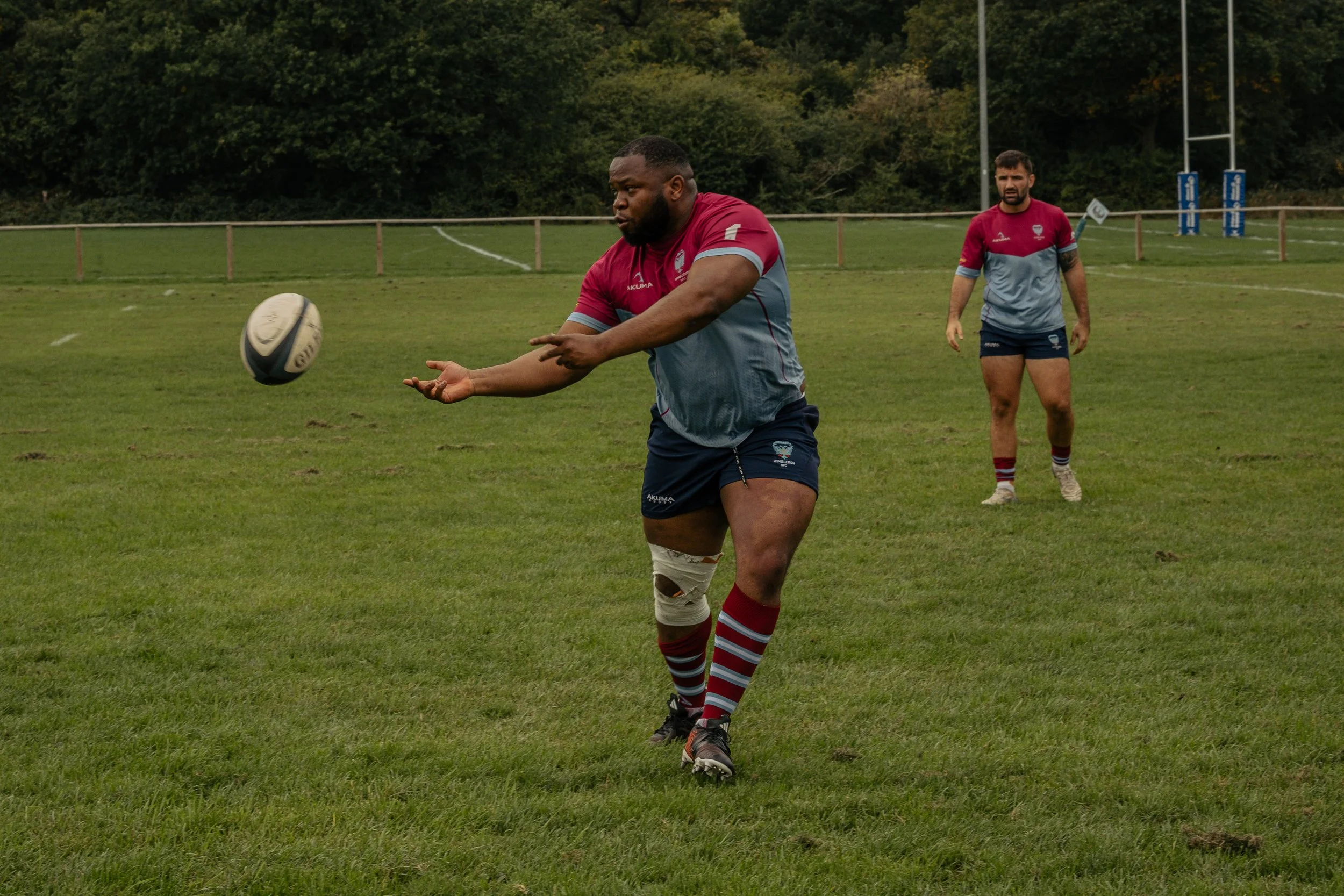 Two rugby players practicing on a grassy field, one is passing a rugby ball while the other watches in the background, both wearing matching maroon and grey uniforms.