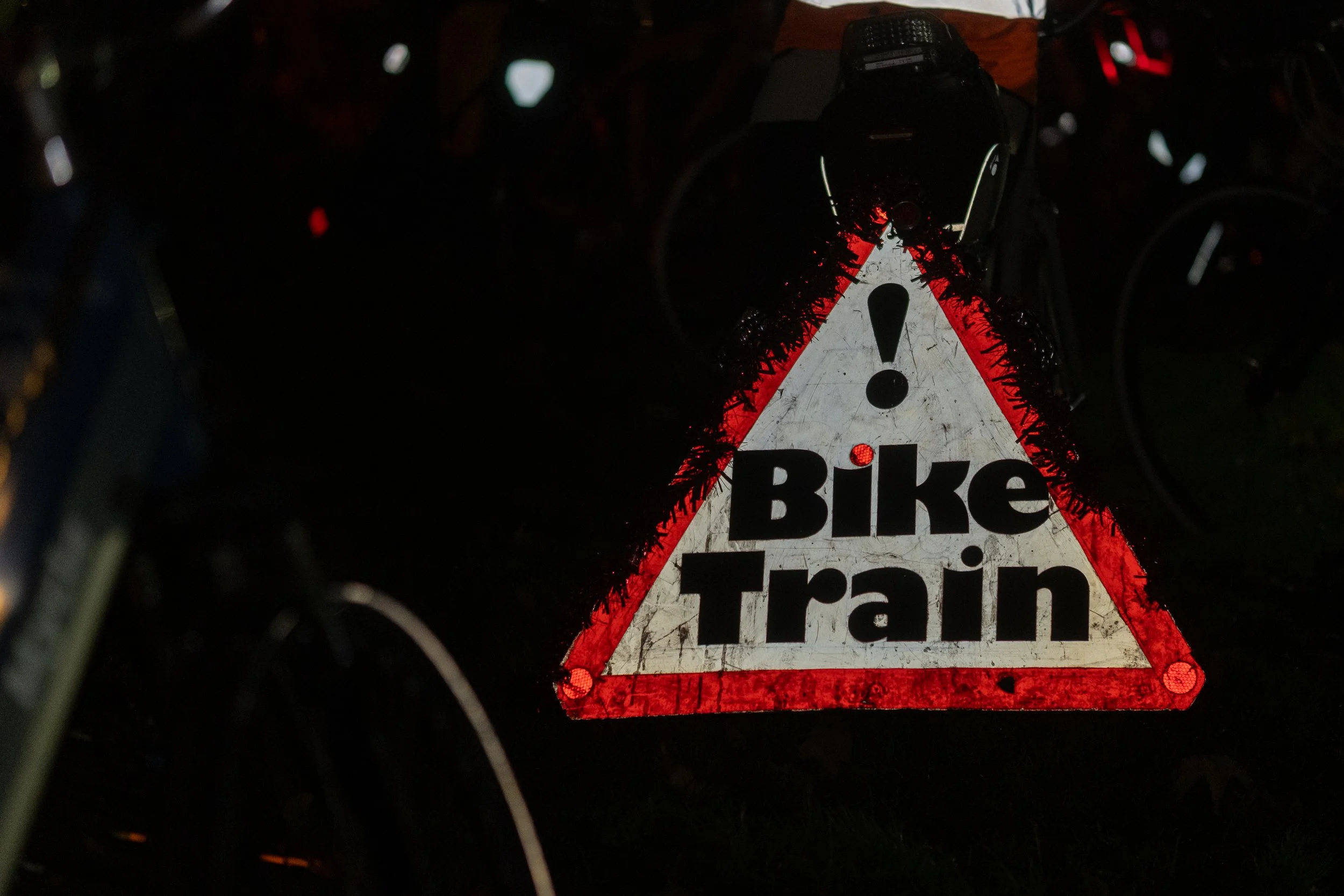 A reflective triangular sign with a red border and the words 'Bike Train' in bold black letters, with a white background and a black exclamation mark at the top, attached to a bicycle at night.