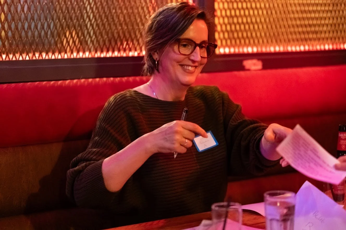 A woman with glasses and short brown hair sitting in a dimly lit restaurant with red and orange lighting. She is smiling, wearing a dark striped sweater, and holding a pen, reaching for a piece of paper handed to her.