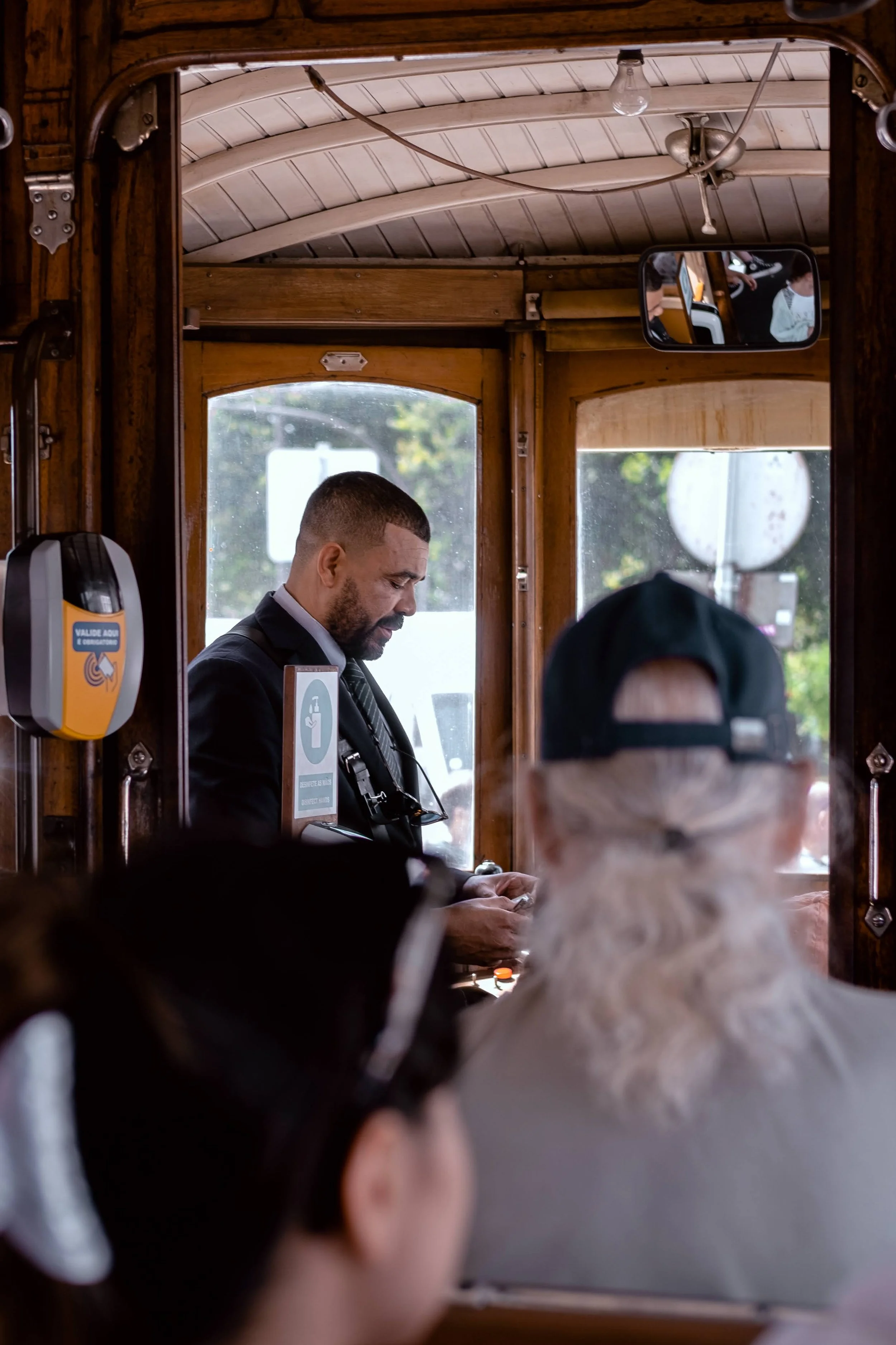 A man in a suit looking at his phone inside a vintage wooden tram with passengers seated in front of him.