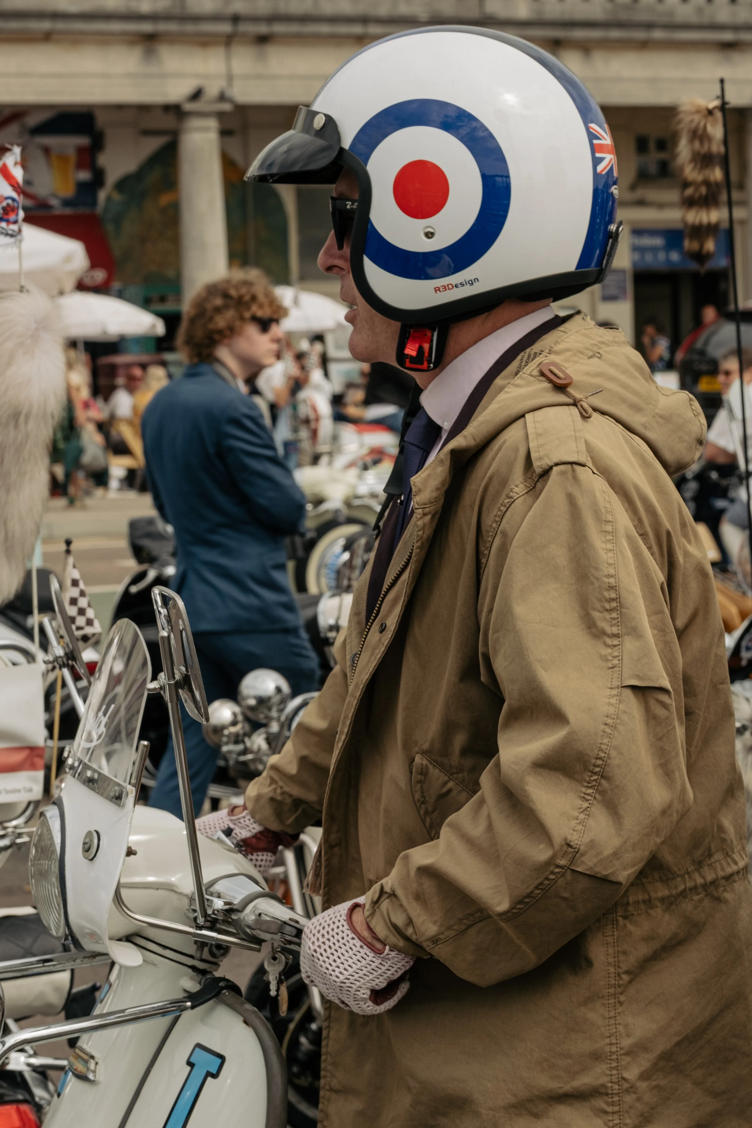 Man in a tan jacket and gloves, wearing a vintage-style motorcycle helmet, standing near a cream-colored scooter at a crowded outdoor event.