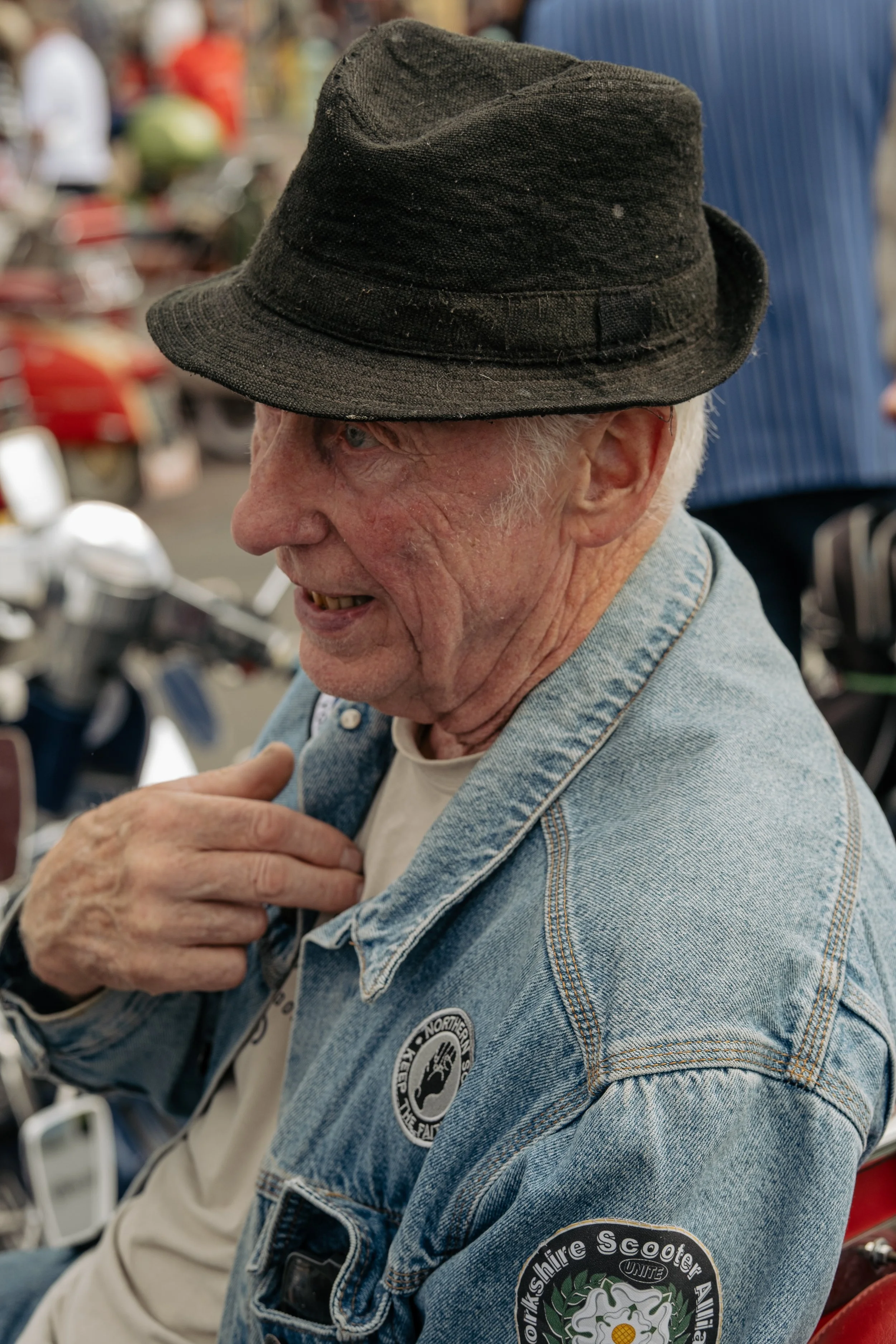 An elderly man wearing a black hat, denim jacket with patches, and a white shirt, sitting near motorcycles at an outdoor event.