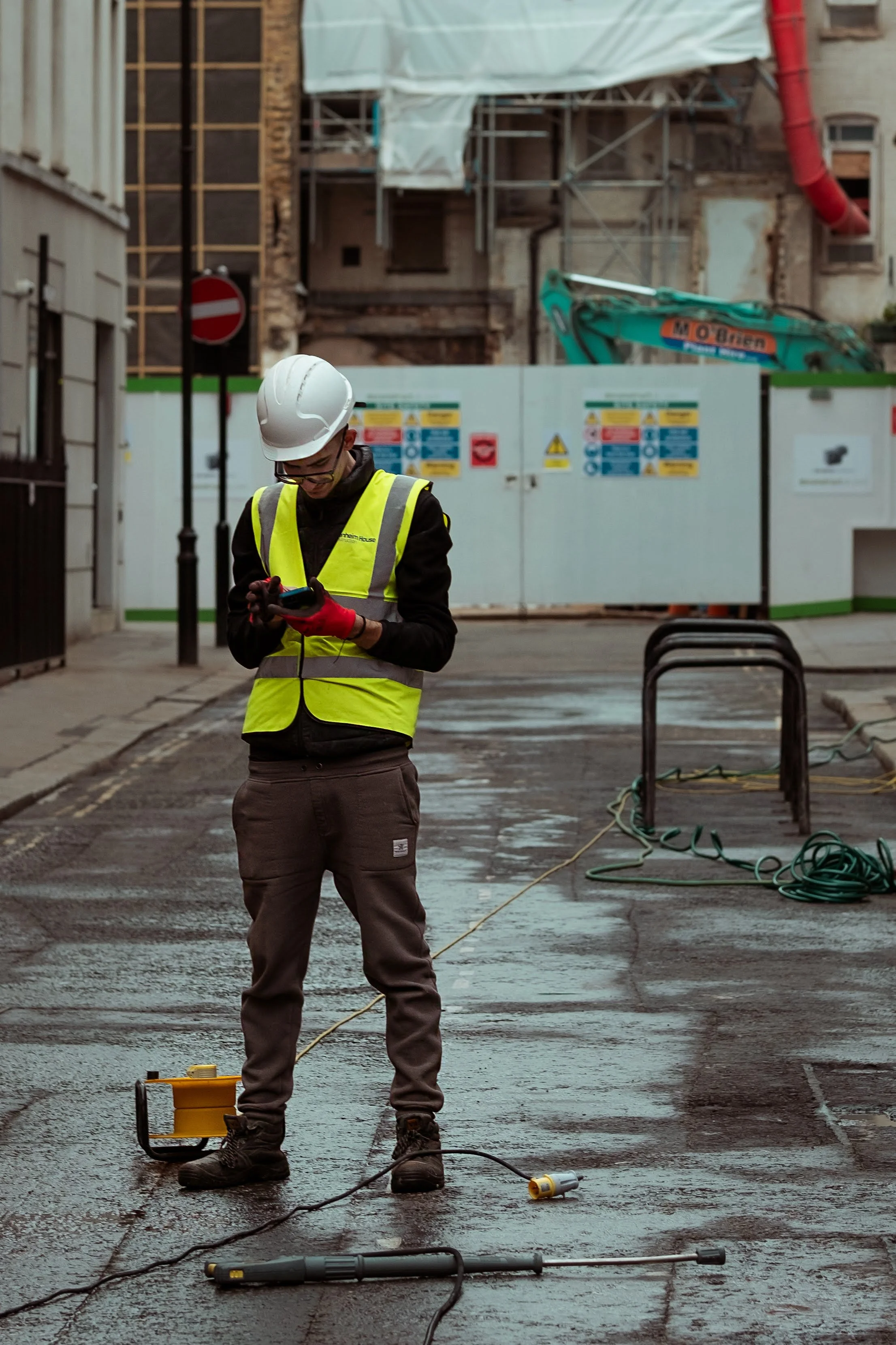 A construction worker in a reflective safety vest, hard hat, and gloves standing on a wet city street, looking down at a device in his hands, with construction equipment and barriers behind him.